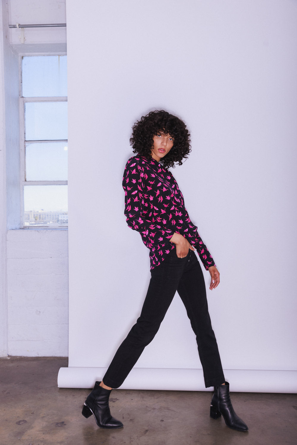 A woman wearing a Catherine Gee Daria French Cuff Silk Pink Crane blouse is predominantly pink with an elegant crane pattern on a black background. It features a classic collar, long sleeves with French cuffs, and a relaxed fit paired with black pants and black ankle boots, creating a stylish and sophisticated look. The plain white background highlights the colors and design of the blouse, making the overall appearance chic and polished, perfect for both casual and formal occasions.
