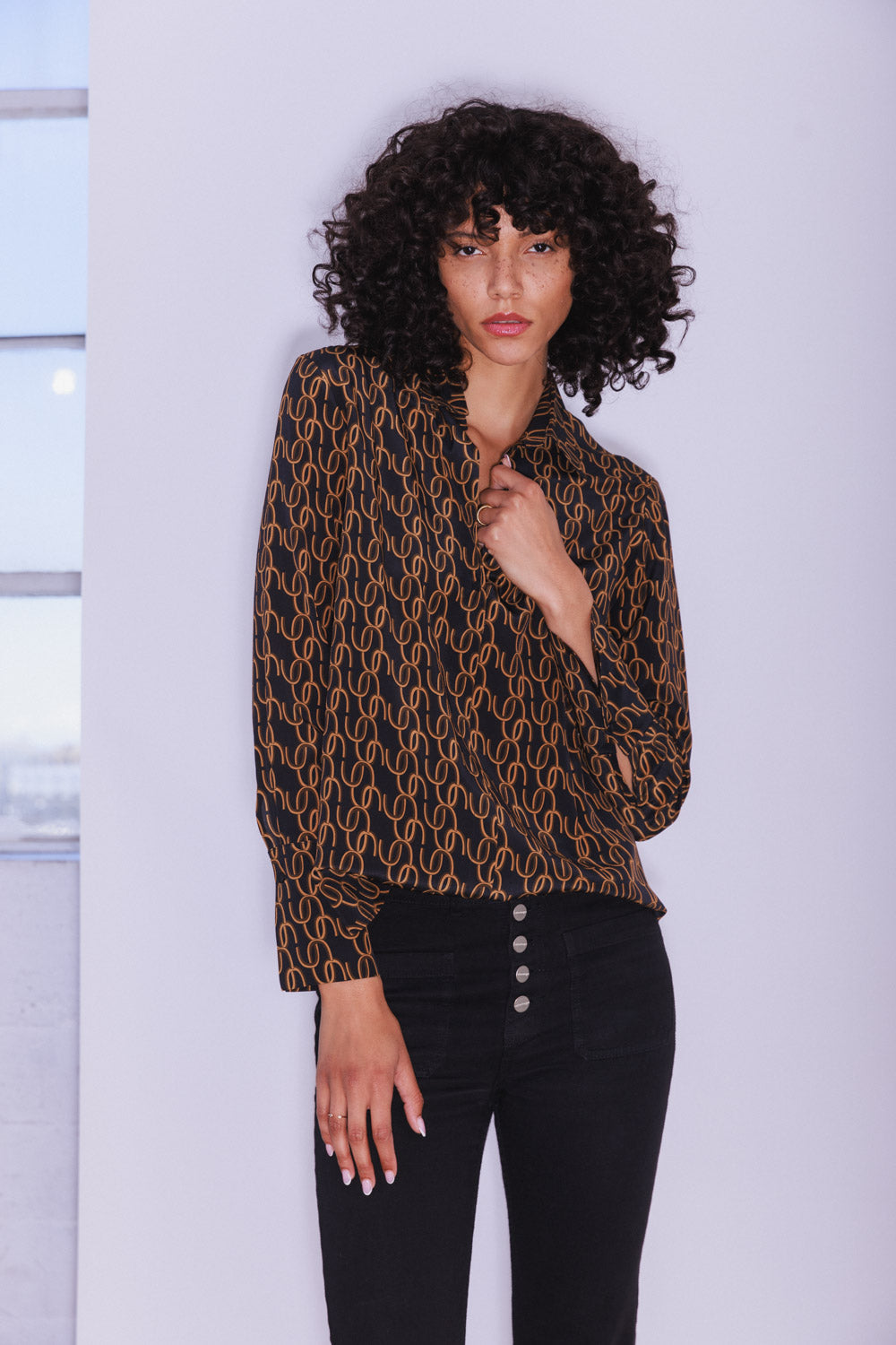 Woman wearing a Catherine Gee Daria French Cuff Silk Blouse CC Print. The blouse is black with a pattern of interlocking gold-colored loops, featuring a straight collar and long sleeves with French cuffs paired with black pants with buttons on the front, standing against a plain, light-colored background with a window beside her.