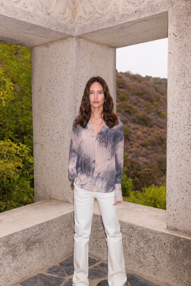 A woman wearing a Catherine Gee Daria French Cuff Silk Smoke blouse, featuring a tie-dye pattern in shades of gray and white. The blouse has a V-neckline and long sleeves with French cuffs paired with white pants, standing against a backdrop of a stone structure and scenic hills with greenery. The luxurious silk fabric and elegant design of the blouse are highlighted, making it a standout piece for a stylish look.