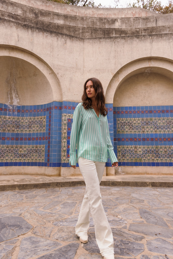 A woman walking in front of a decorative wall with blue and patterned tiles wearing a Catherine Gee Daria French Cuff Silk Mint Green Stripe blouse paired with white pants and white shoes. The background features an architectural structure with arches and a stone floor.