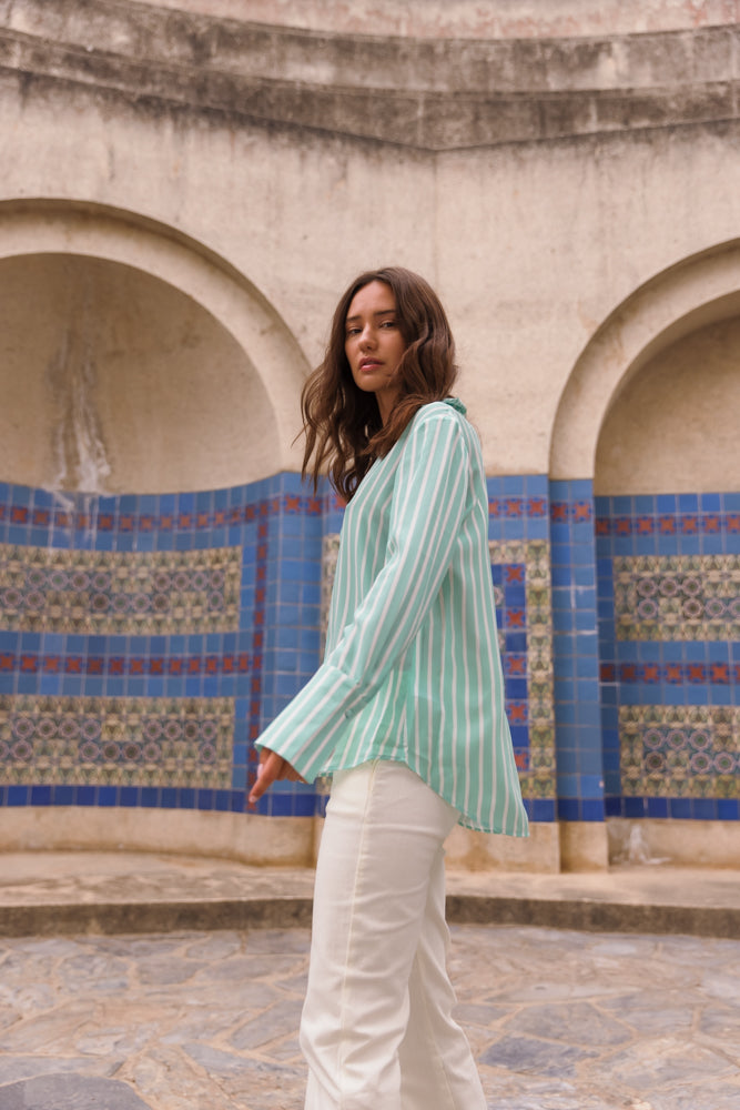 A woman standing in front of a decorative tiled wall with arches features an architectural structure with arches and a stone floor. She wears a Catherine Gee Daria French Cuff Silk Mint Green Stripe blouse and white pants. The background features intricate blue and patterned tiles, adding an artistic and cultural element to the scene.