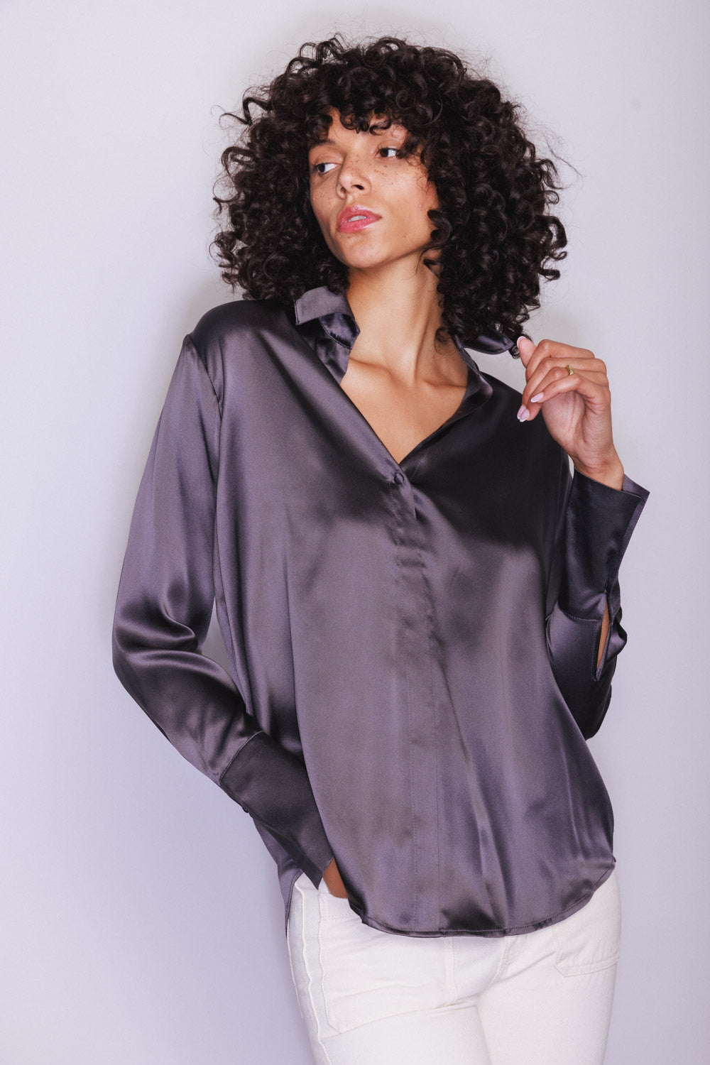 A woman wearing a Catherine Gee Daria French Cuff Silk Pewter blouse features a sleek, pewter-colored silk fabric with a subtle metallic sheen, has a V-neckline, and long sleeves with French cuffs, which add a touch of elegance. The blouse has a relaxed fit and is paired with white pants, creating a sophisticated and polished look. The overall design and luxurious material make this blouse a standout piece for any wardrobe.