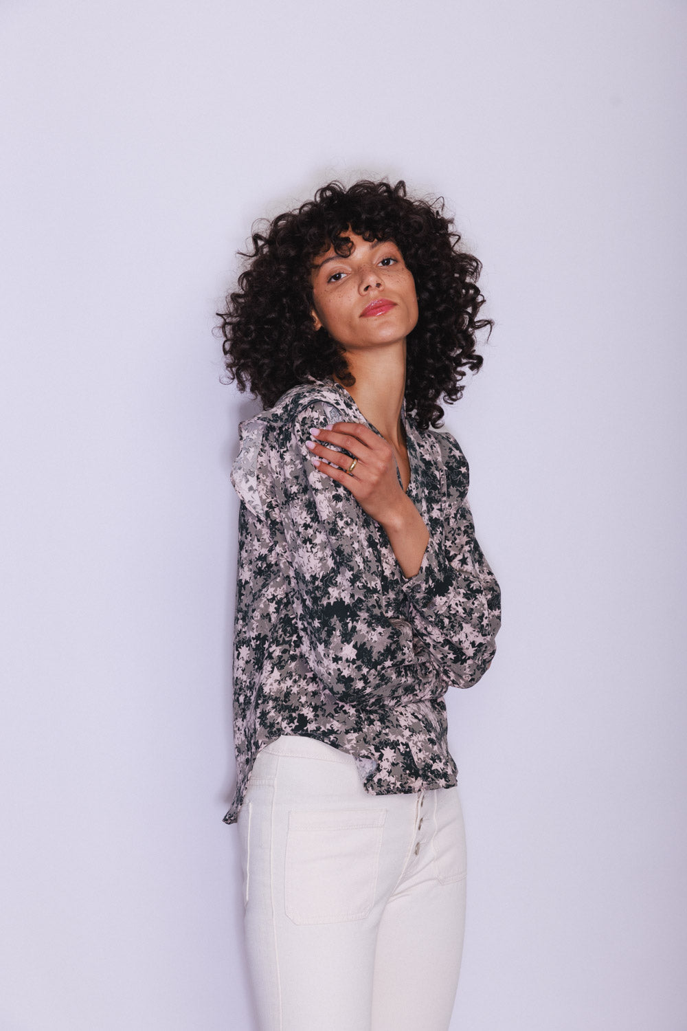 A woman wearing a Catherine Gee Franka Starshower blouse features a sophisticated floral pattern in shades of black, white, and grey. It has a loose fit with long sleeves and a slightly ruffled collar, giving it an elegant touch paired with white pants, creating a chic contrast. The background is plain and light-colored, emphasizing the blouse's stylish design.