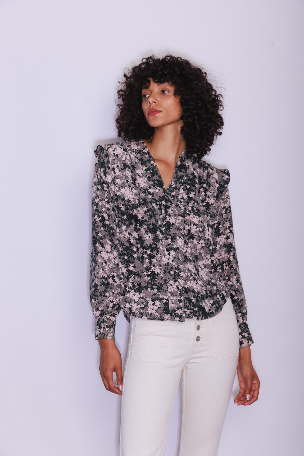 A woman wearing a Catherine Gee Franka Starshower blouse that features a dark floral pattern in shades of black and pink. This creates an elegant and eye-catching design, which she pairs with high-waisted white pants. The blouse has a V-neckline and long sleeves, contributing to its sophisticated look and highlighting its intricate details. Overall, the outfit exudes a stylish vibe.