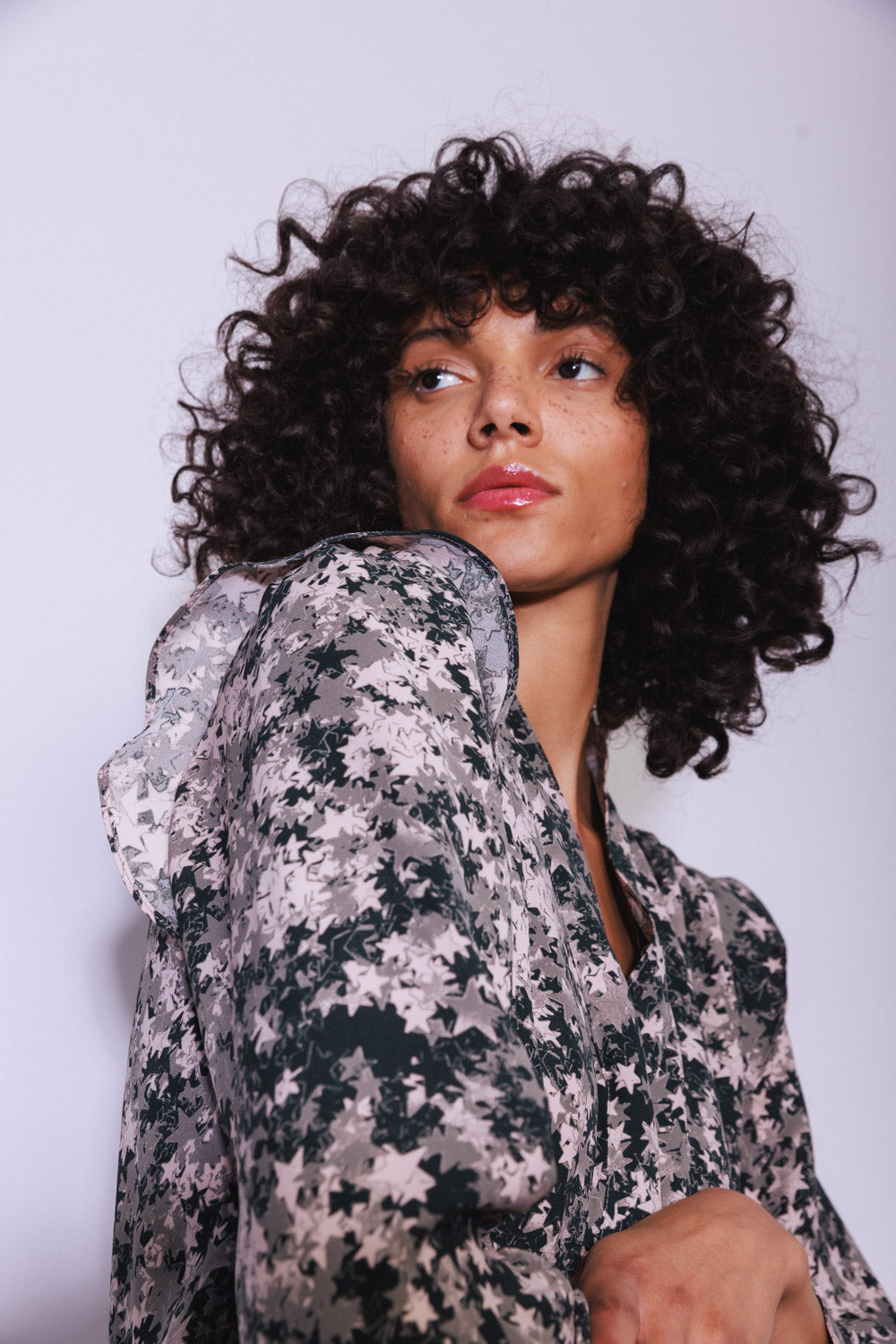 A woman with curly hair posed with one arm slightly raised wearing a Catherine Gee Franka Starshower blouse that features a delicate, star-patterned design in shades of black, white, and grey. The fabric appears lightweight and semi-sheer, with a loose, flowing fit highlighting the blouse's sleeve and pattern.