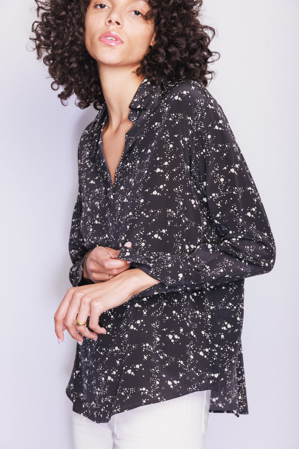 A woman wearing a Catherine Gee Daria French Cuff Silk Dot Heart Blouse features a black color with a white heart-shaped and splatter dot pattern. The blouse has a V-neck and long sleeves with French cuffs paired with white pants showcasing the design and fit of the blouse.