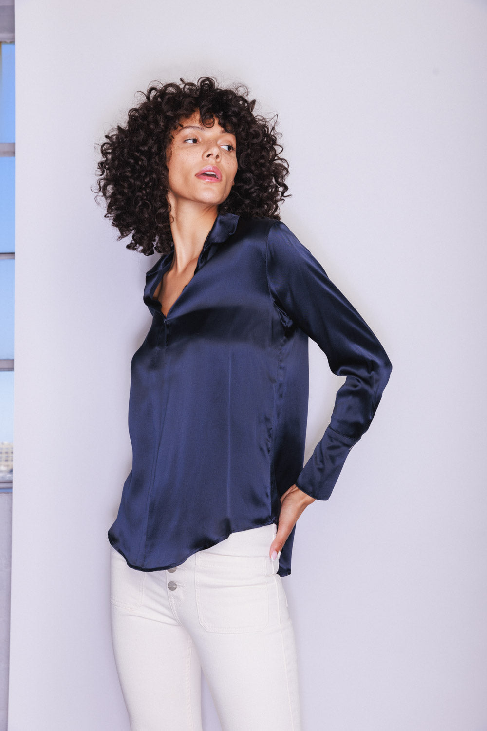 A woman wearing a Catherine Gee Daria French Cuff Silk-Midnight Navy blouse features a smooth, shiny texture and a relaxed fit with long sleeves and a V-neckline paired with white pants and posing with one hand in a pocket on a plain white background with a hint of a window on the right side.