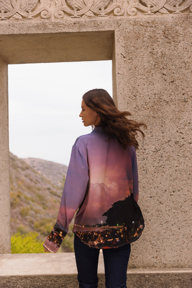 A woman with long hair standing with her back to the camera, wearing a Catherine Gee Daria French Cuff Silk Bali blouse features a scenic print with a sunset or sunrise over a landscape, possibly depicting a coastal or mountainous region. She is framed by a stone window or archway, with a natural landscape visible in the background highlighting the detailed and vibrant design of the blouse, making it interesting for showcasing fashion and scenic art combined in clothing.