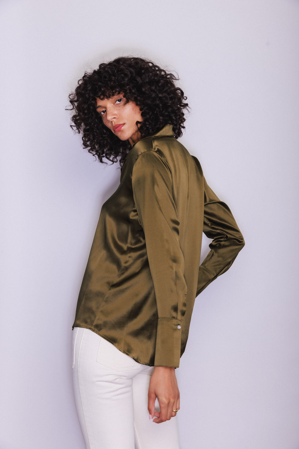 A woman wearing a Catherine Gee Daria French Cuff Silk Blouse Army Green. The blouse has a smooth, shiny texture and long sleeves with French cuffs. She is also wearing white pants and is posed with one hand on the hip, showcasing the side profile of the blouse. The background is plain and light-colored, highlighting the details of the blouse.