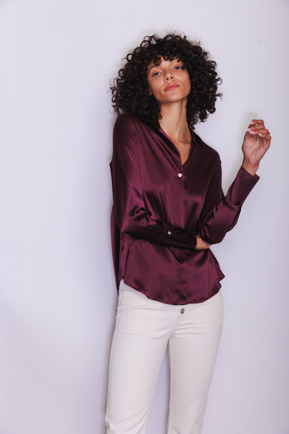 A woman wearing a Catherine Gee Daria French Cuff Silk Wine blouse is a rich, deep wine color with a silky texture, featuring a V-neckline and French cuffs paired with light-colored pants, which contrast beautifully with the dark blouse. The overall look is elegant and sophisticated, highlighting the luxurious fabric and stylish design of the blouse.