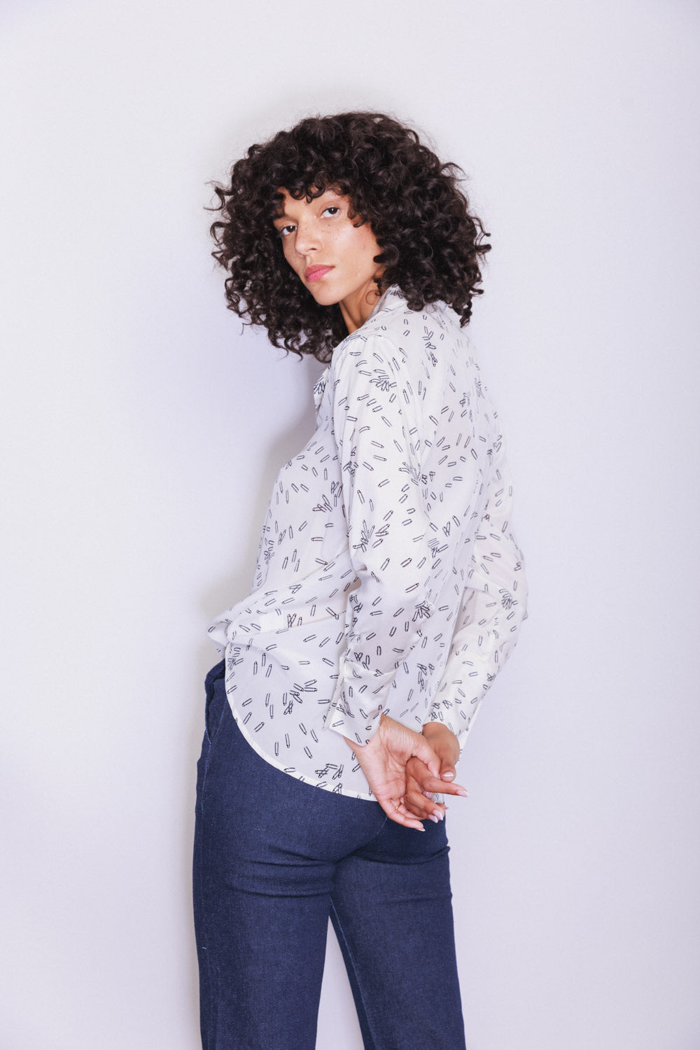 A woman wearing a Catherine Gee Daria French Cuff Silk Pin blouse features a white silk fabric adorned with a delicate pattern of small black shapes, includes a straight collar, long sleeves with French cuffs, and covered buttons, adding a touch of sophistication. The blouse is paired with dark blue pants, creating a polished and stylish look. The overall outfit is perfect for both professional and casual settings, making it a versatile addition to any wardrobe.