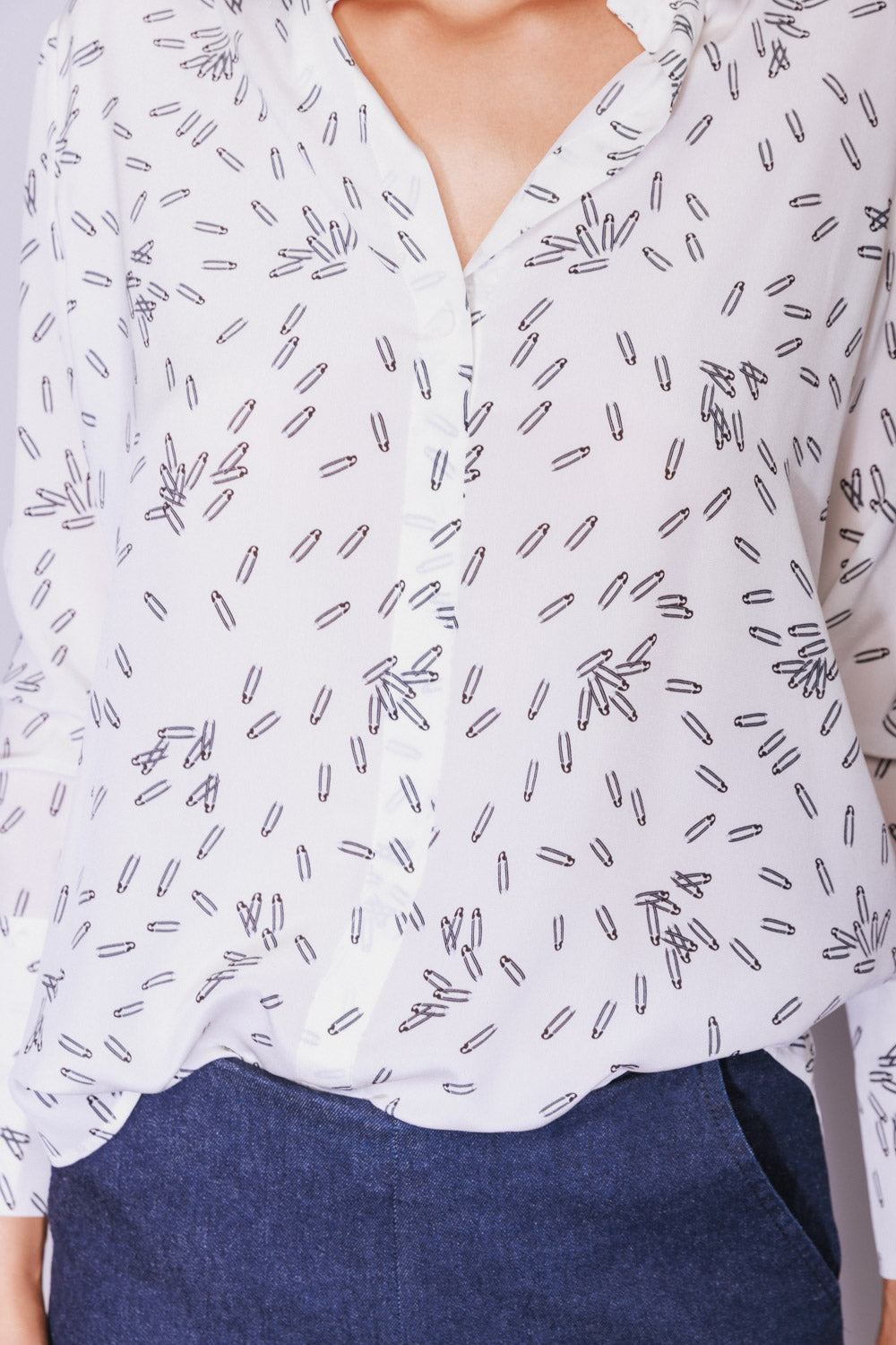 A close-up view of a Catherine Gee Daria French Cuff Silk Pin blouse features white with a pattern of small, black, elongated shapes scattered throughout the fabric, a V-neckline with a slight ruffle detail along the edge, and a button-down front paired with dark blue pants. The blouse appears to be made of a lightweight, silky material. The design is casual yet elegant, making it a versatile piece for various occasions.