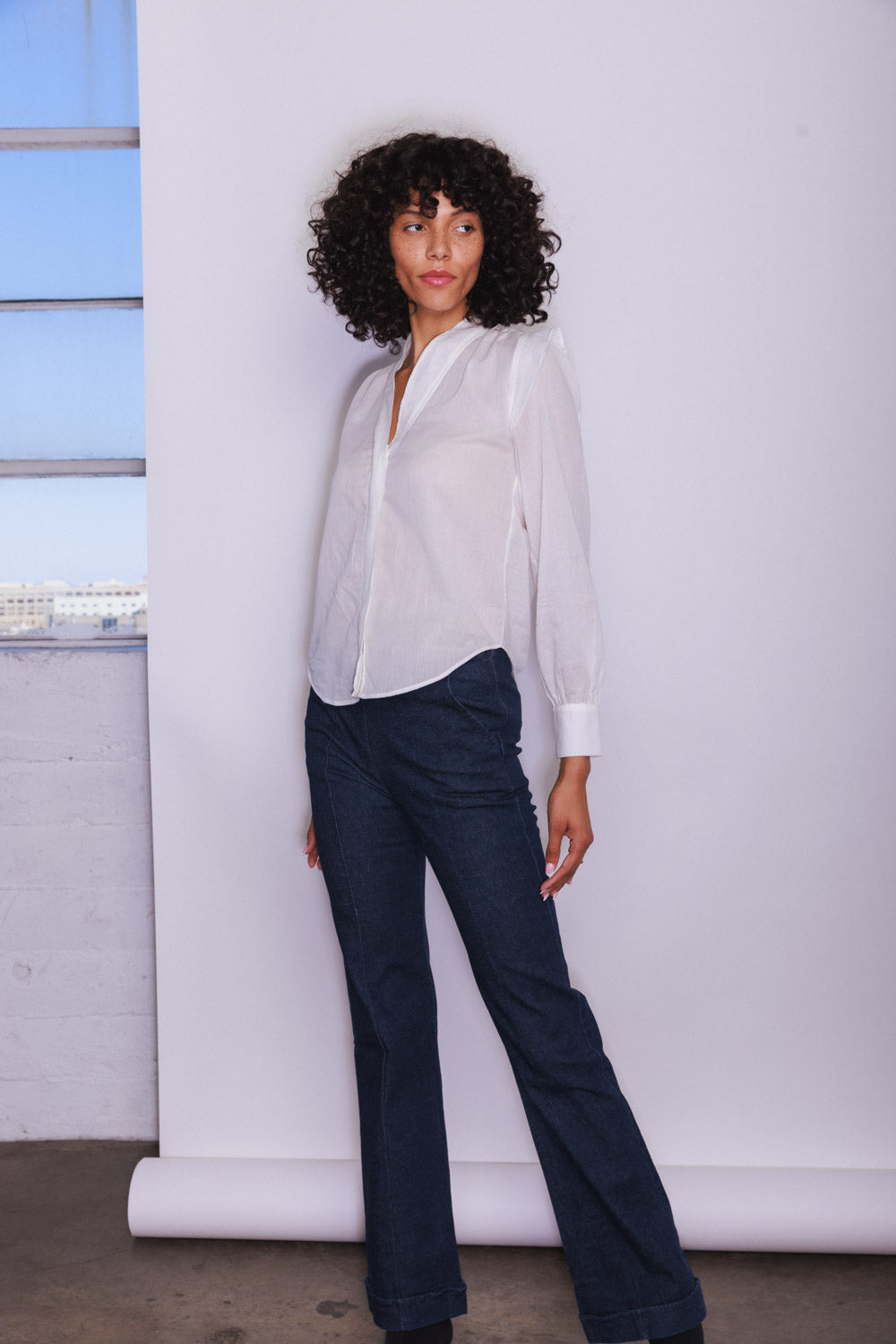 A woman wearing a Catherine Gee Franka White Blouse paired with dark blue jeans. The blouse is a classic white button-down with a slightly loose fit, long sleeves, and a collar. She is standing against a plain white backdrop, with a window to the left side of the image, allowing natural light to illuminate the scene. The outfit is simple yet elegant, highlighting the timeless style of the white blouse.