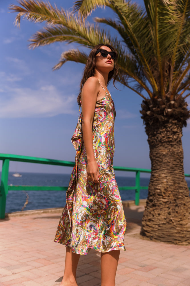 A woman standing outdoors near a palm tree and a green railing, with the ocean and a clear blue sky in the background wearing a Catherine Gee Emma Paint Swirl Slip Dress. The dress features a vibrant, abstract swirl pattern with a mix of bright and bold colors, and has thin spaghetti straps, and a graceful V-neckline. This setting enhances the dress's summery and breezy feel, making it perfect for warm, coastal environments.