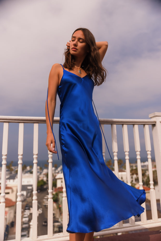A woman wearing a Catherine Gee Emma Mykonos Blue Slip Dress standing on a balcony with a white railing, and the background includes a view of a coastal town with buildings and a partly cloudy sky. The dress is a vibrant blue color with a silky texture, featuring thin shoulder straps and a flowing, elegant design that creates a striking and visually appealing contrast.