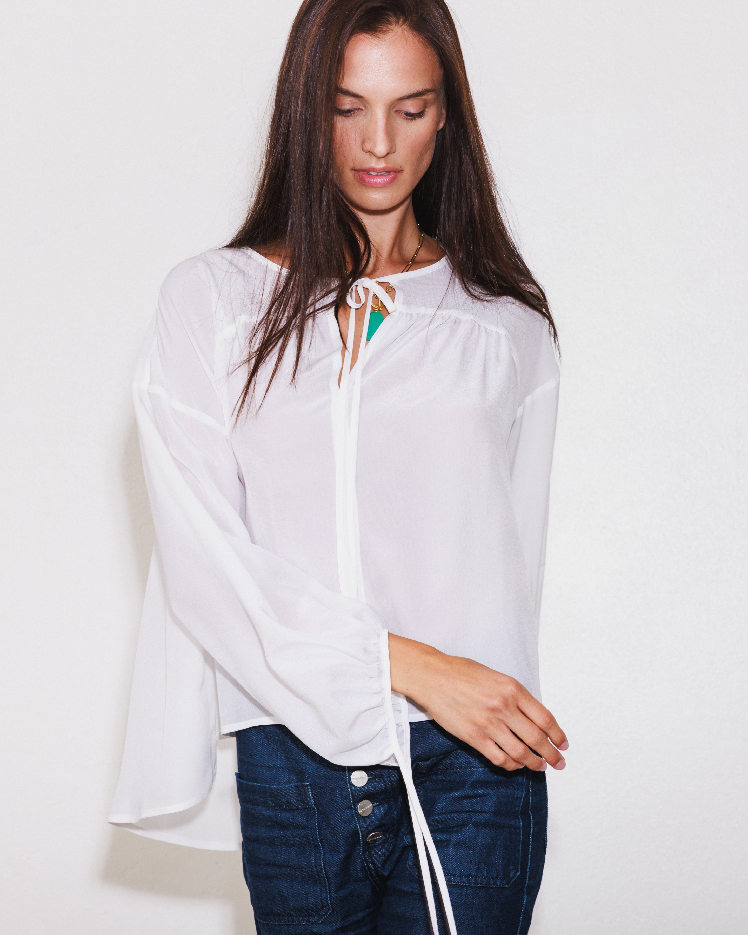 A woman wearing a Catherine Gee Frances White Blouse. This blouse has a loose, flowing design with long sleeves that gather at the wrists and tie with delicate strings. The neckline features a subtle tie detail, adding a touch of sophistication. The fabric is lightweight and slightly sheer, creating an elegant and airy look. She pairs the blouse with dark blue jeans adorned with multiple buttons, combining casual comfort with a sophisticated style.