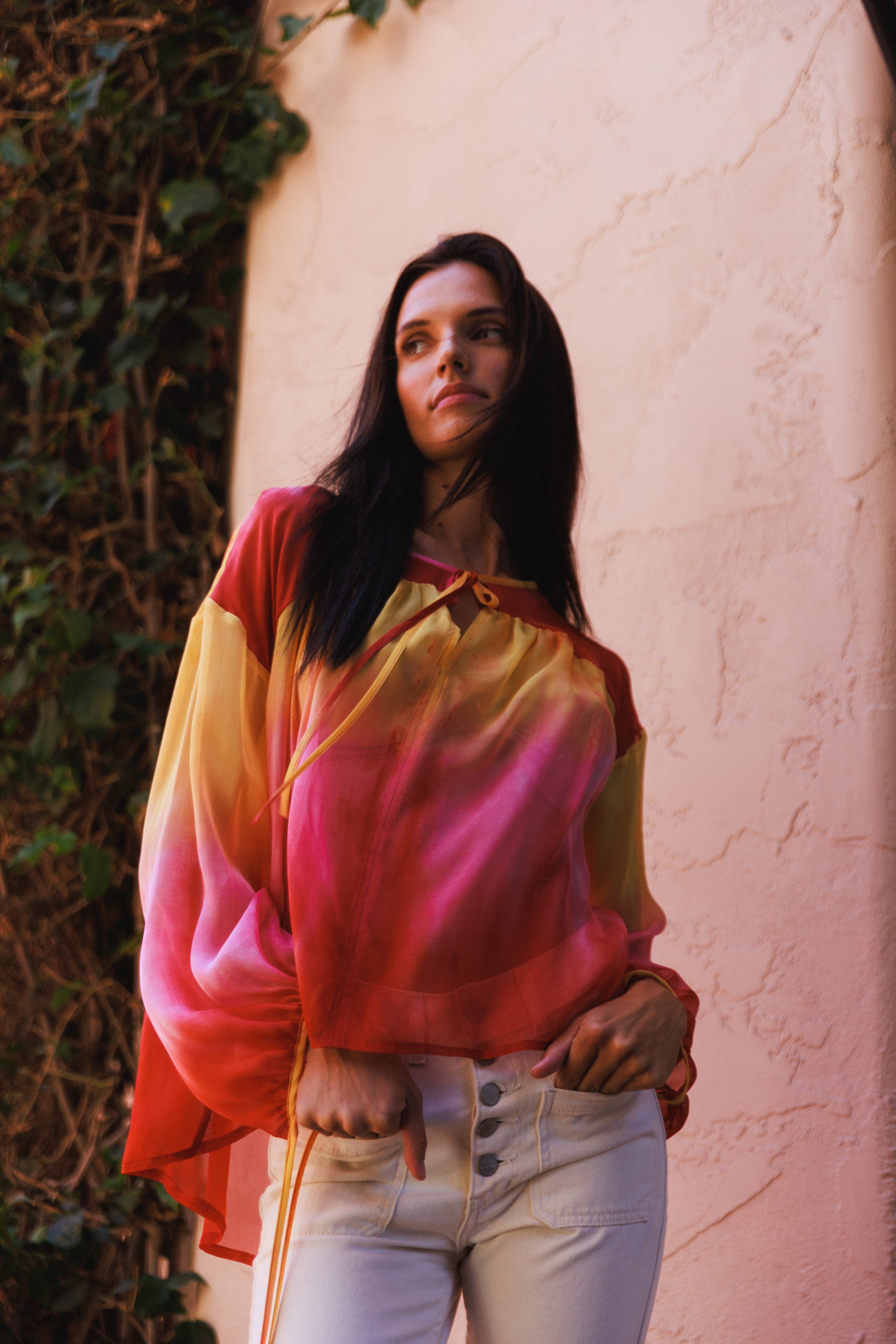 A woman wearing the Catherine Gee Frances Santa Barbara Sunset blouse features a stunning gradient from yellow at the top to pink and red at the bottom, reminiscent of a sunset. It has long, flowing sleeves and a loose, airy fit paired with light-colored, high-waisted pants, the ensemble exudes an effortless elegance. The background includes a textured wall and greenery, adding a natural touch to the vibrant outfit.