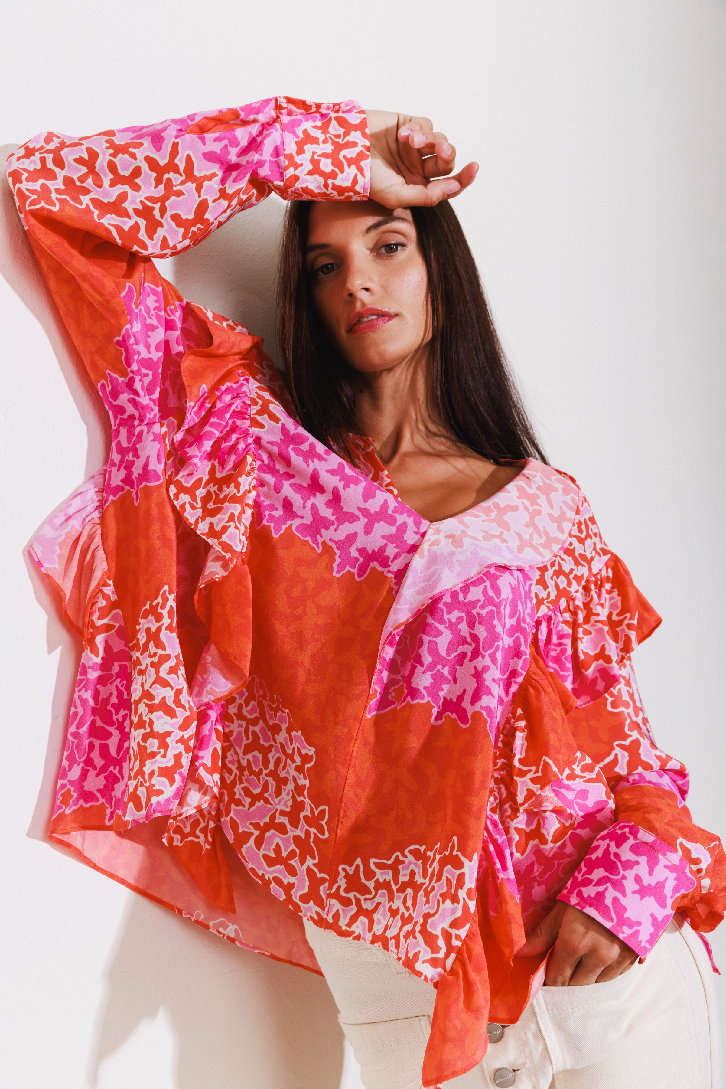 A woman wearing a Catherine Gee Bella - Mariposa blouse features a vibrant design with shades of red, pink, and orange, adorned with abstract floral patterns. The blouse has a loose fit with ruffled details on the sleeves and body. She is posing with one arm raised and the other hand resting on the hip, against a plain white background.