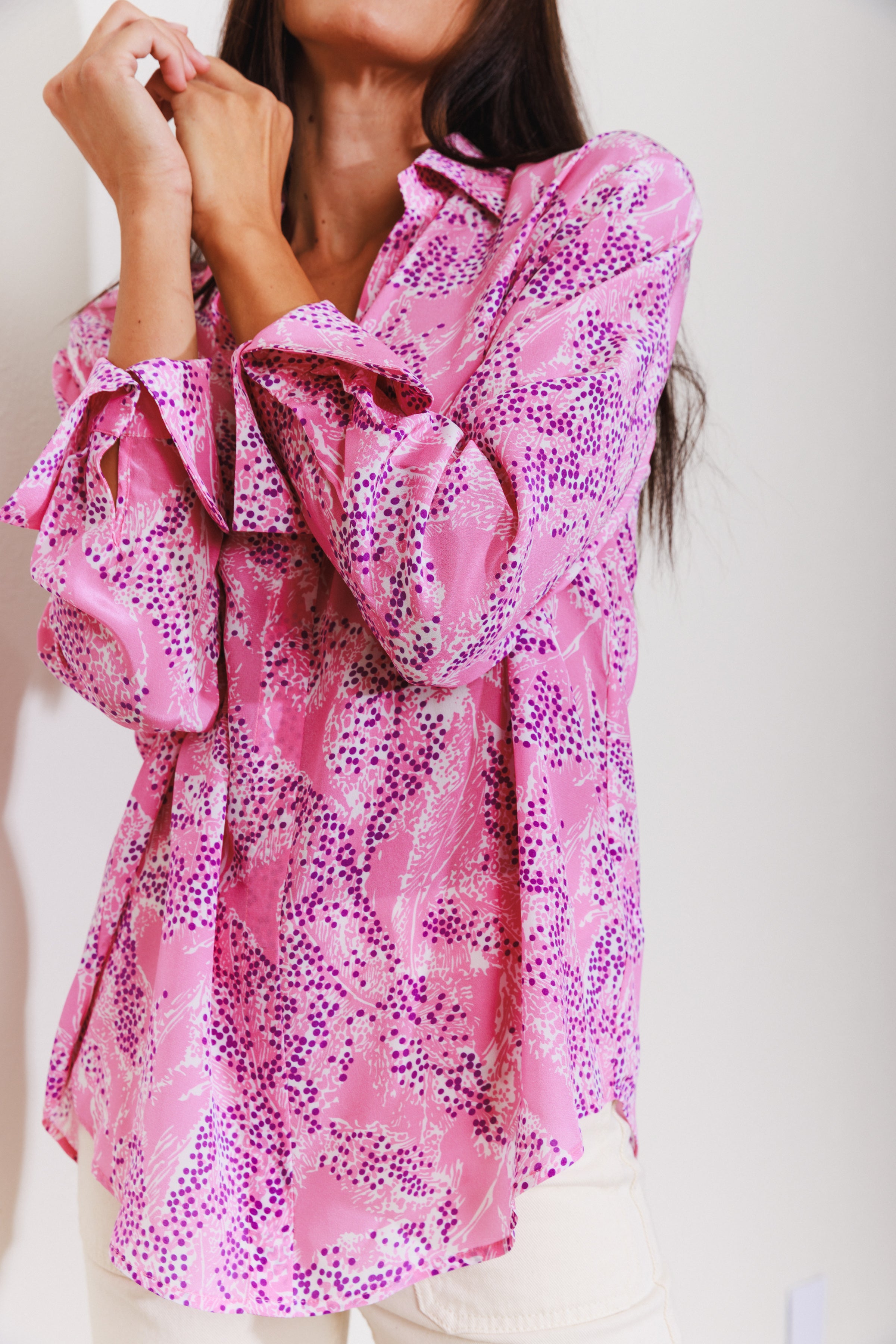 A woman wearing a Catherine Gee Daria French Cuff Silk Pink Sea blouse features a vibrant pattern with shades of pink, purple, and white, resembling abstract sea elements. It has French cuffs and a loose, flowing fit, enhancing the elegance of the silky fabric. She's posing with hands near the face, highlighting the blouse's design and texture, perfect for making a statement in any setting.