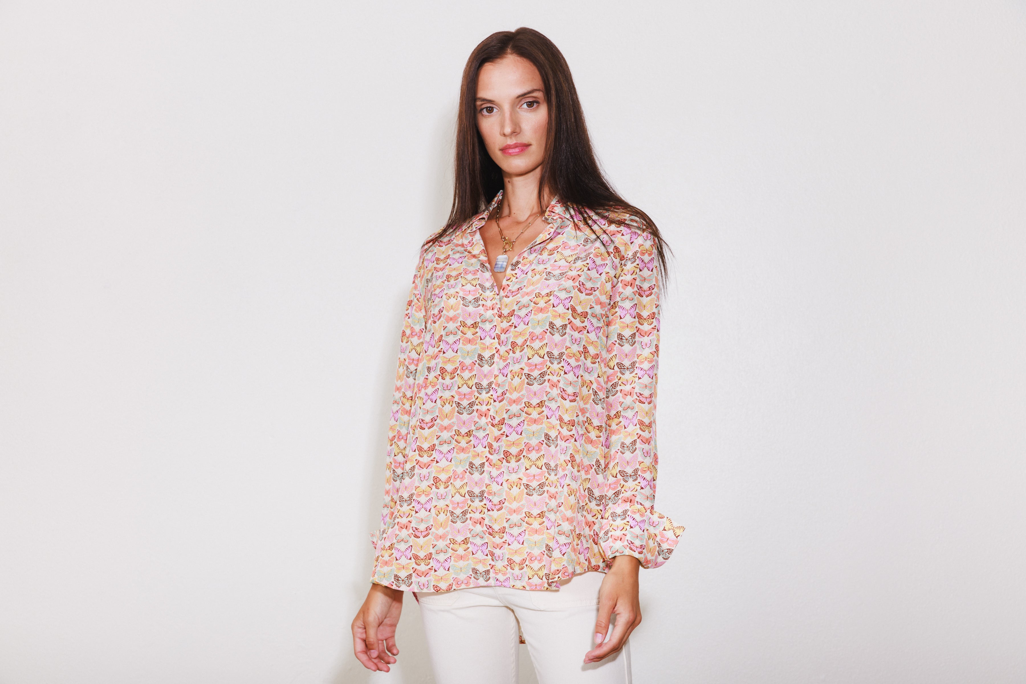 A woman wearing a Catherine Gee Daria French Cuff Silk blouse features a colorful butterfly pattern on a light background, with long sleeves and a relaxed fit. She is also wearing light-colored pants and a necklace with a pendant on a plain white background.