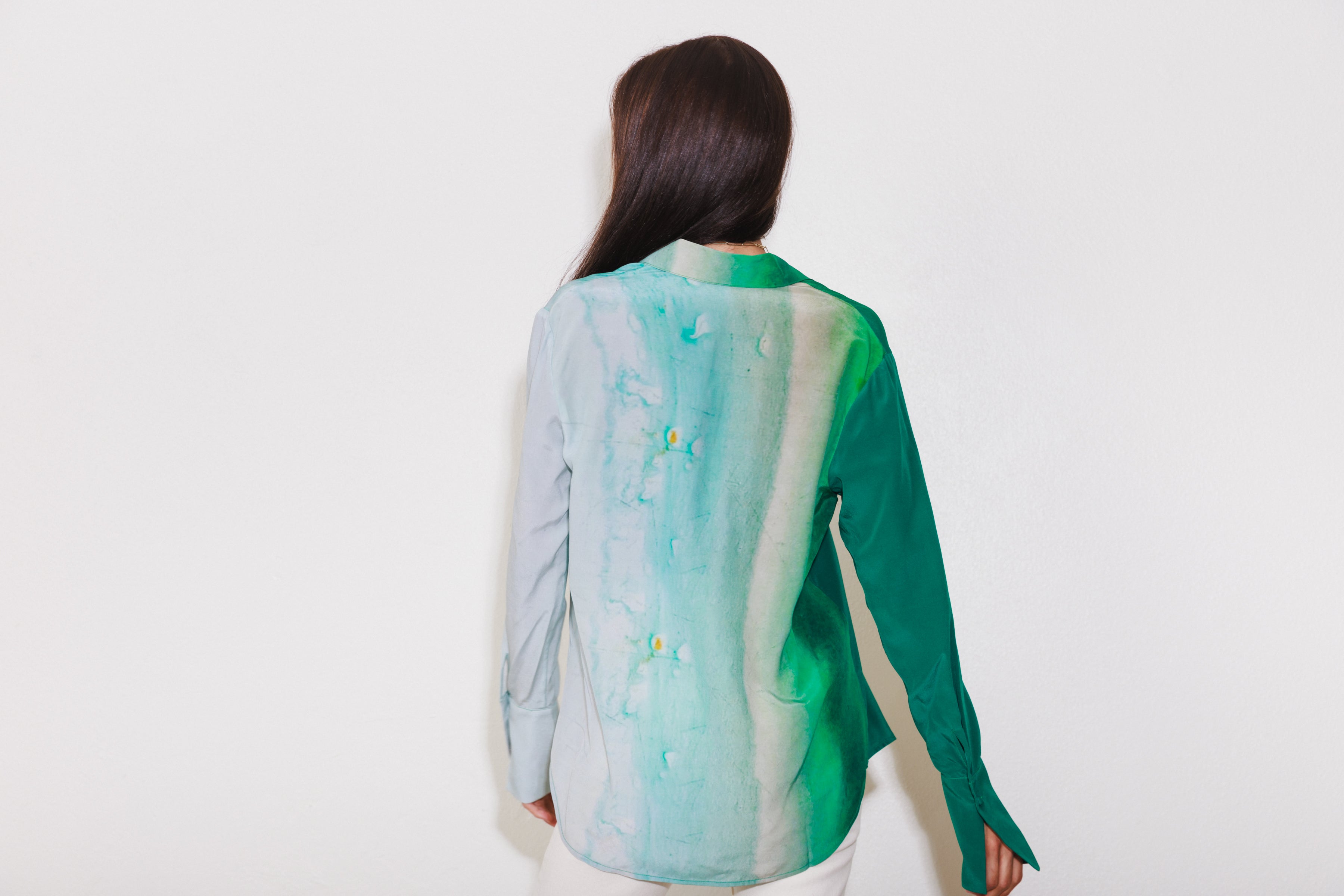 A woman with long dark hair is shown from the back standing against a plain white background wearing a Catherine Gee Daria French Cuff Silk-King Wash blouse features a unique design with a gradient of green shades, transitioning from a light mint green on the left side to a darker green on the right side. The blouse has a smooth, silky texture and long sleeves with French cuffs.