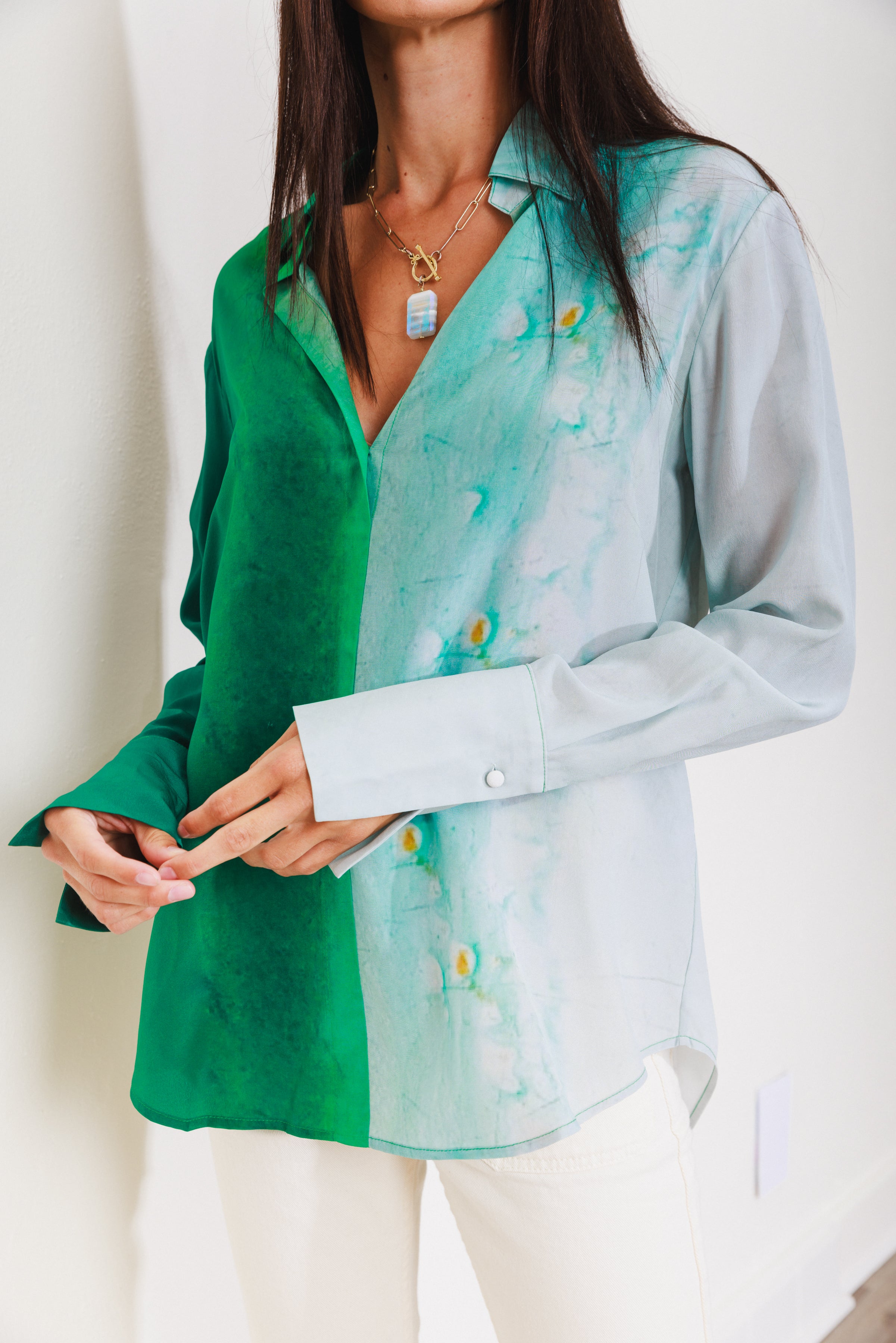 A woman wearing a Catherine Gee Daria French Cuff Silk-King Wash blouse features a unique design with a green color on the left side and a light blue color with a marbled pattern on the right side. It has a V-neckline and long sleeves with French cuffs. She also wears white pants and a necklace with a pendant.