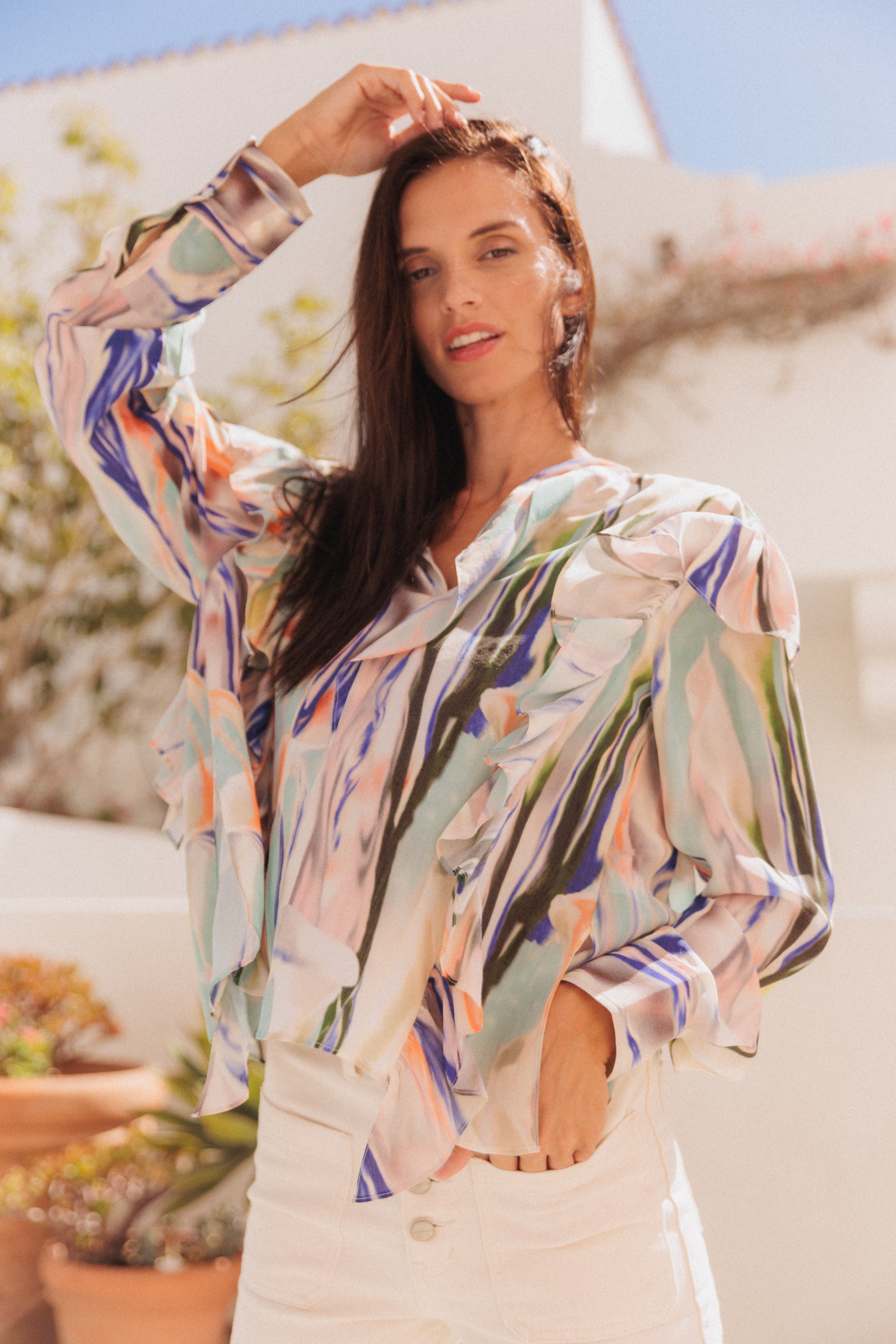 A woman is wearing a Catherine Gee Bella Blouse - Abstract Orchid features a colorful, abstract floral design with shades of blue, green, orange, and white paired with white pants and is standing outdoors with a blurred background that includes some plants and a white building. The woman has one hand raised to her head and the other one inside the left side pocket of the pants. The image highlights the vibrant and artistic design of the blouse, making it the focal point.