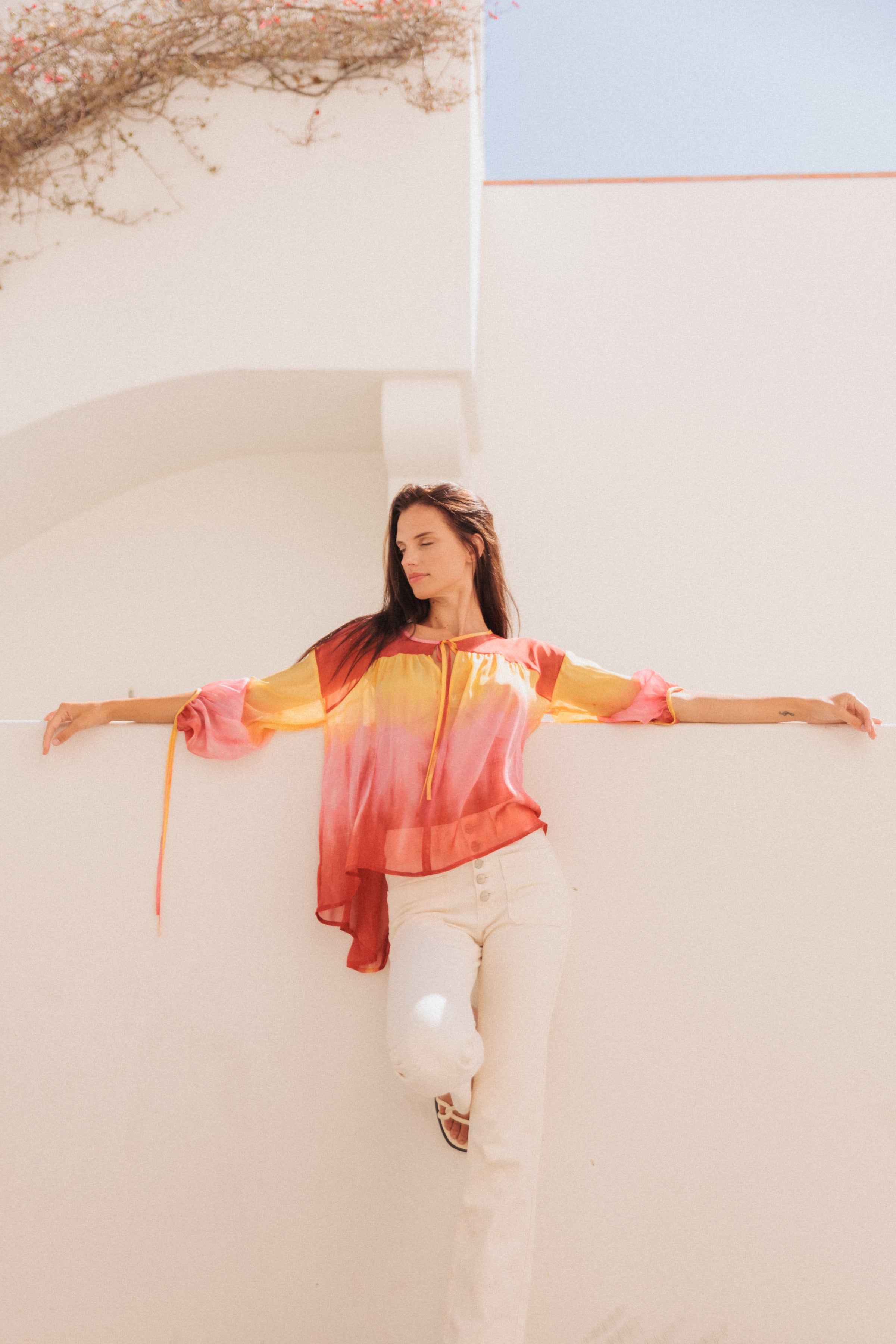 A woman wearing the Catherine Gee Frances Santa Barbara Sunset blouse features a vibrant gradient of colors that transitions from yellow at the top to pink and red at the bottom. The blouse has long sleeves and a loose, flowing fit. She stands against a white wall with her arms outstretched, creating a striking contrast between the colorful blouse and the clear blue sky background and some foliage visible at the top.