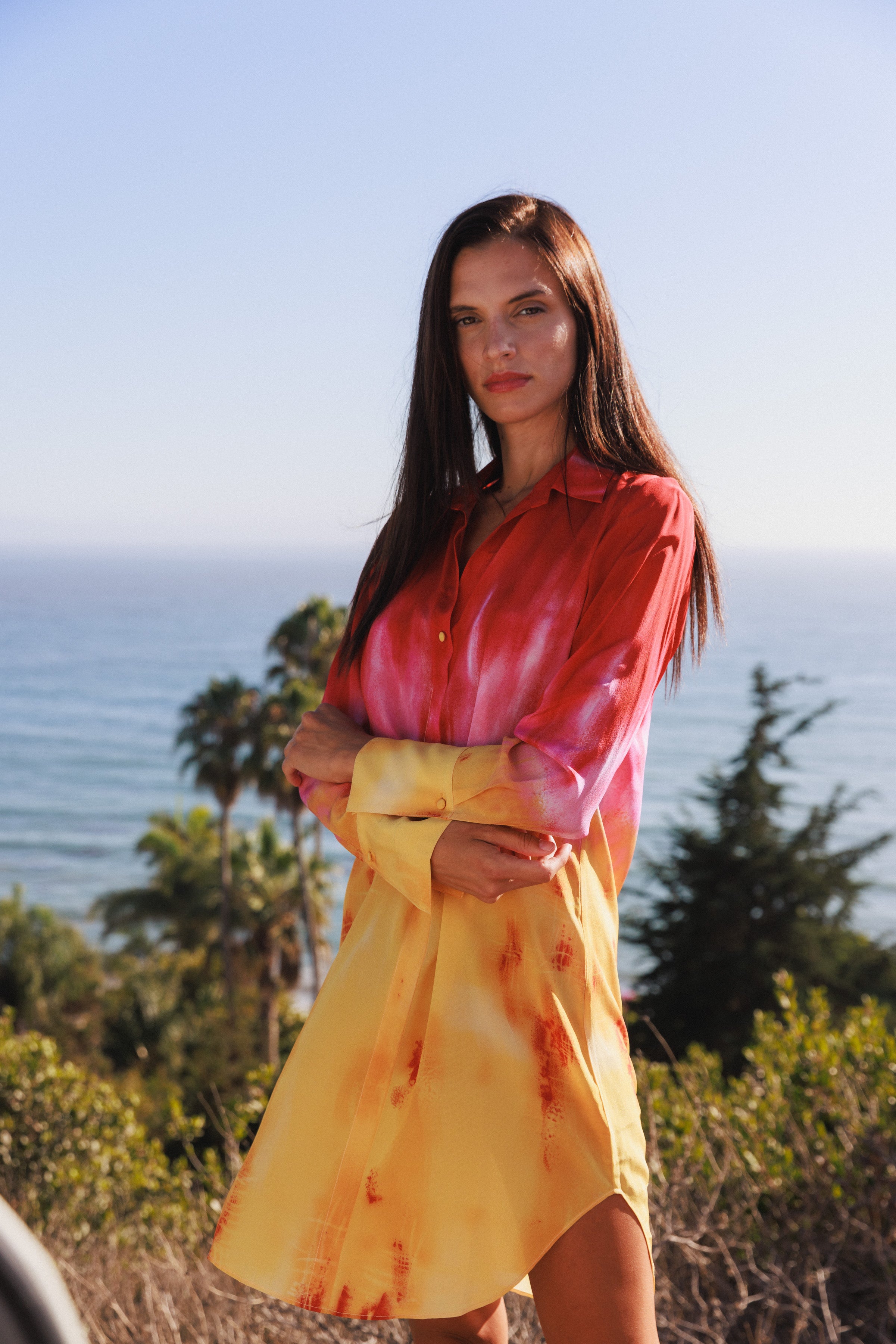 A woman wearing a Catherine Gee Daria Silk Shirt Santa Barbara Sunset Dress. This vibrant dress features a stunning tie-dye pattern with red and pink hues on the top transitioning to yellow at the bottom. It has a relaxed fit, long sleeves, and a collar, embodying a perfect blend of casual elegance. She is standing with a scenic ocean view, palm trees, and greenery in the background, enhancing the dress's summery appeal.