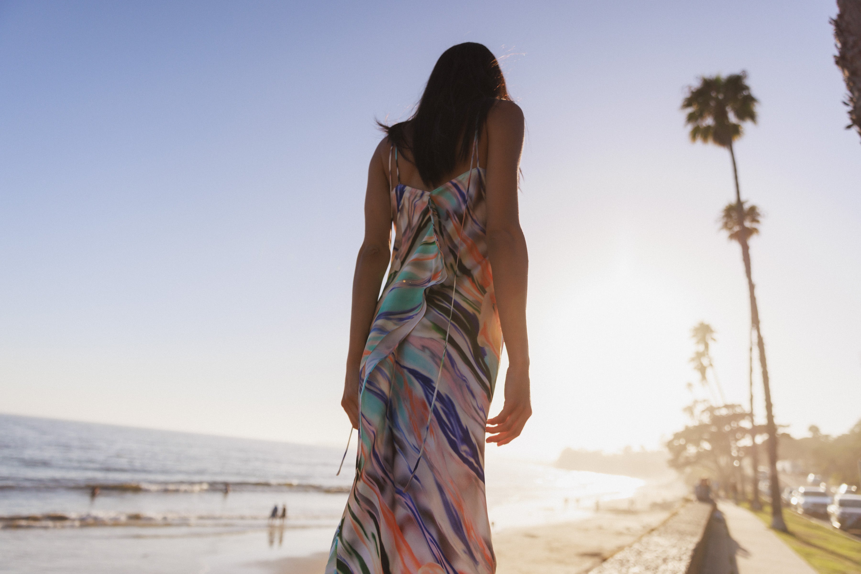 A woman walking along a beachside during sunset, wearing a Catherine Gee Emma Abstract Orchid Slip dress features a vibrant, colorful, abstract pattern with shades of blue, purple, green, orange, beige, and white. The beach, ocean, and palm trees are visible in the background, creating a serene and picturesque scene. The warm glow of the setting sun enhances the dress's vibrant colors, making it a stunning piece for any occasion.