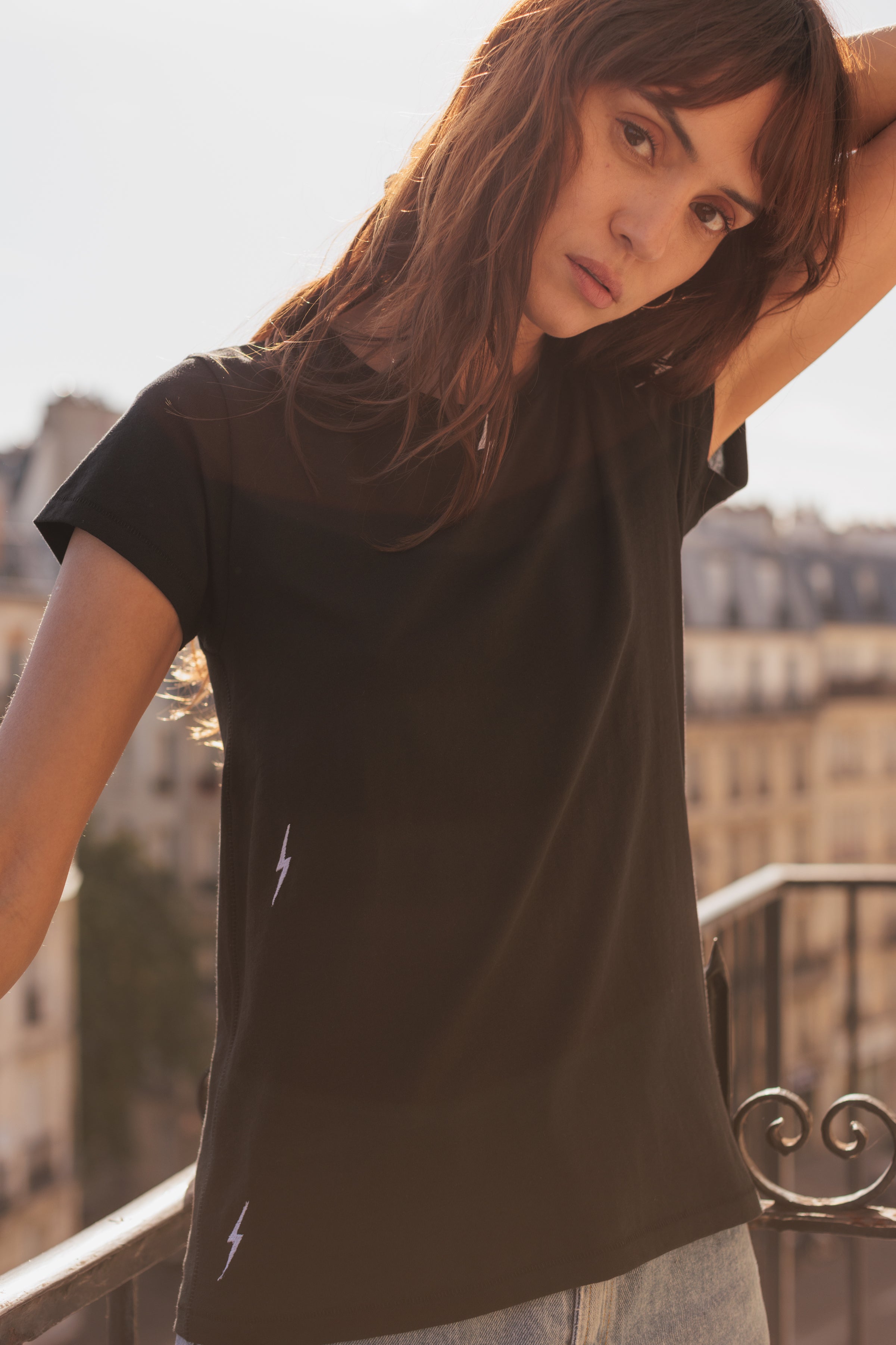 A woman standing on a balcony with an urban backdrop wearing a Catherine Gee Embroidered Cotton Bolt black T-Shirt features short sleeves and a classic crew neck design with a small, white lightning bolt embroidery near the bottom left side of the shirt, adding a unique and edgy touch highlighting the casual yet stylish vibe of this versatile tee.