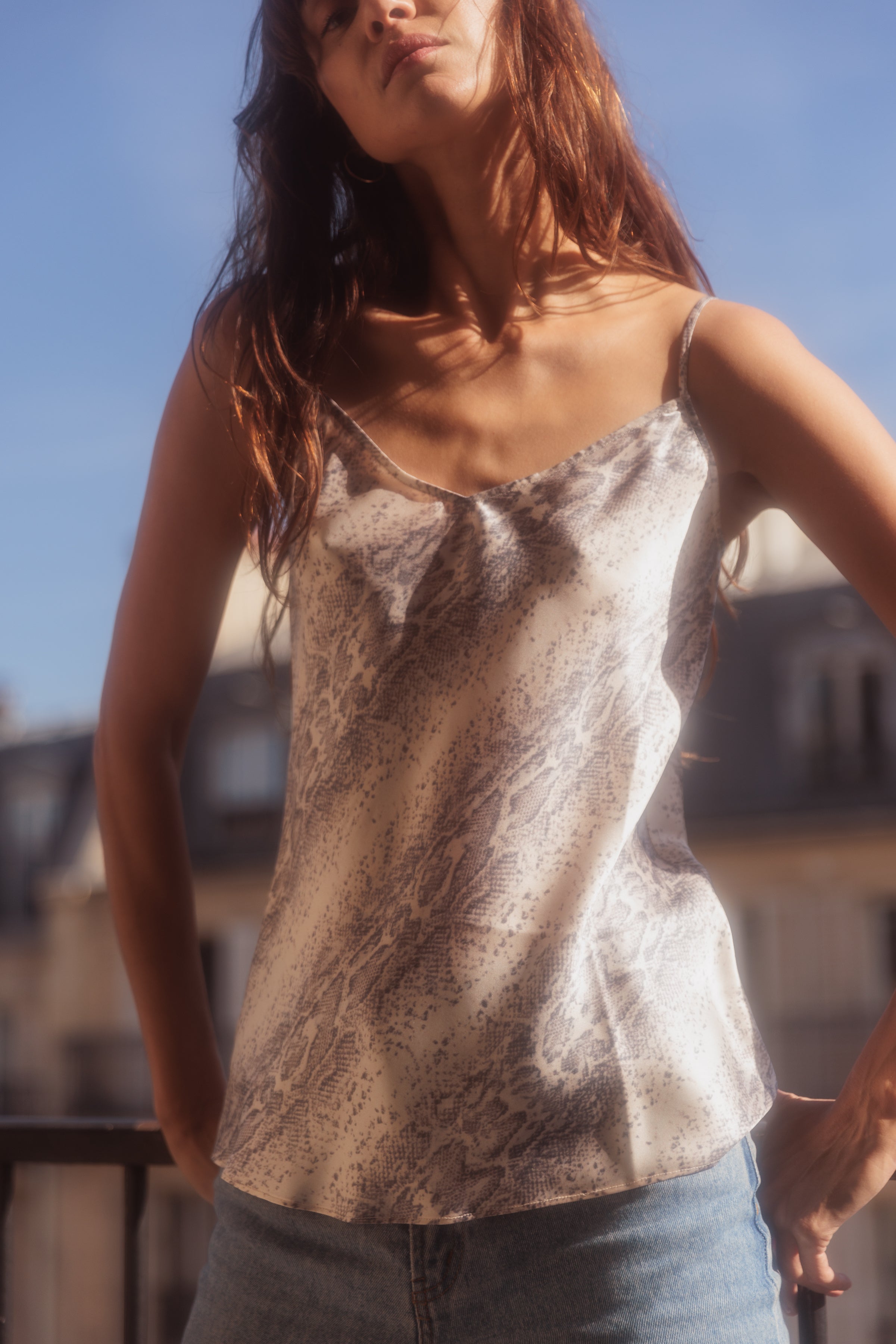 A woman wearing a Catherine Gee Basic Cami - Sedona Snake. The cami features a delicate snake print pattern and thin straps paired with blue jeans and is standing in front of a blurred background of buildings under a clear blue sky.