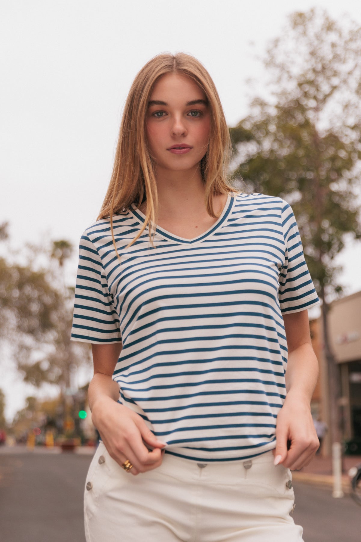 A woman wearing the Catherine Gee Jen Relaxed Shortsleeve - Navy Stripe tee features a classic navy and white horizontal stripes, a V-neckline, and short sleeves paired with white shorts with button details on the sides. The background includes an outdoor setting with trees and buildings, suggesting a casual, everyday look. The combination of the tee and shorts creates a stylish and relaxed outfit, perfect for a day out.