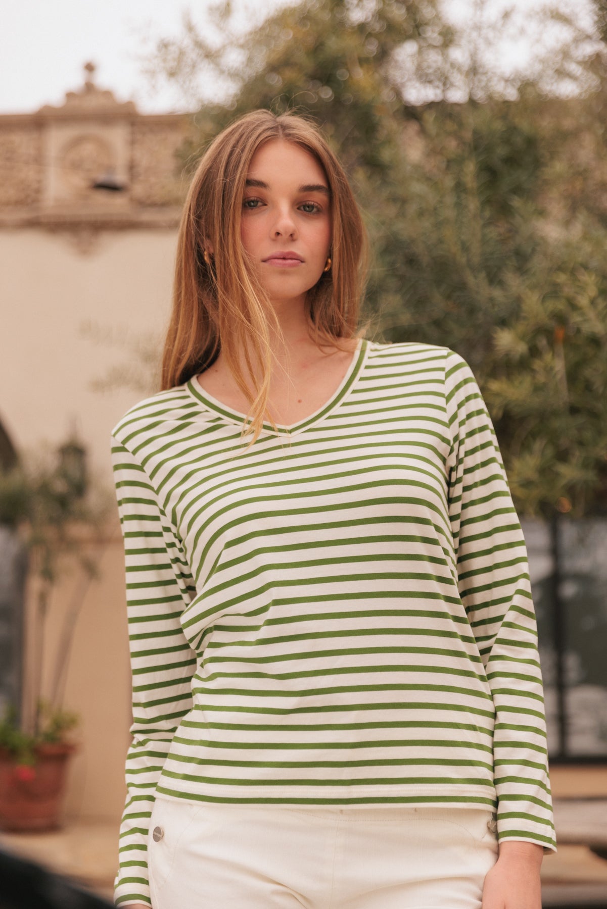 A woman wearing a Catherine Gee Jen Relaxed Longsleeve - Green Stripe tee features horizontal green and white stripes, a V-neckline, and long sleeves paired with white pants with button details. The background includes some greenery and a building with architectural details, suggesting an outdoor setting. The image highlights the casual and stylish nature of the tee, making it relevant for fashion enthusiasts.