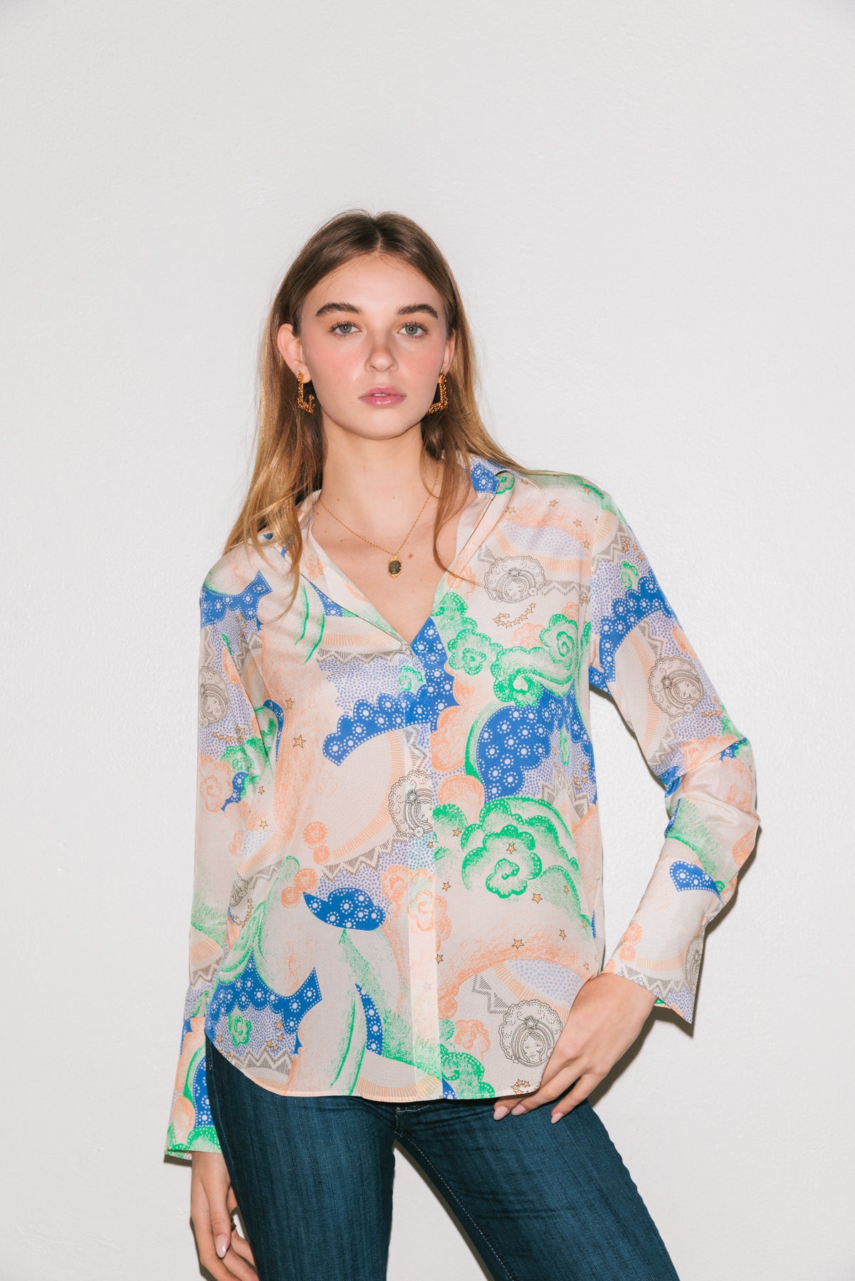 A woman wearing a Catherine Gee Daria French Cuff Silk Siena blouse features an intricate and colorful pattern with shades of blue, green, and peach on a light background. It has a V-neckline and long sleeves with French cuffs. Paired with dark blue jeans. The outfit displays both elegance and casual charm. The luxurious silk fabric and detailed design make the blouse a standout piece, perfect for various occasions.