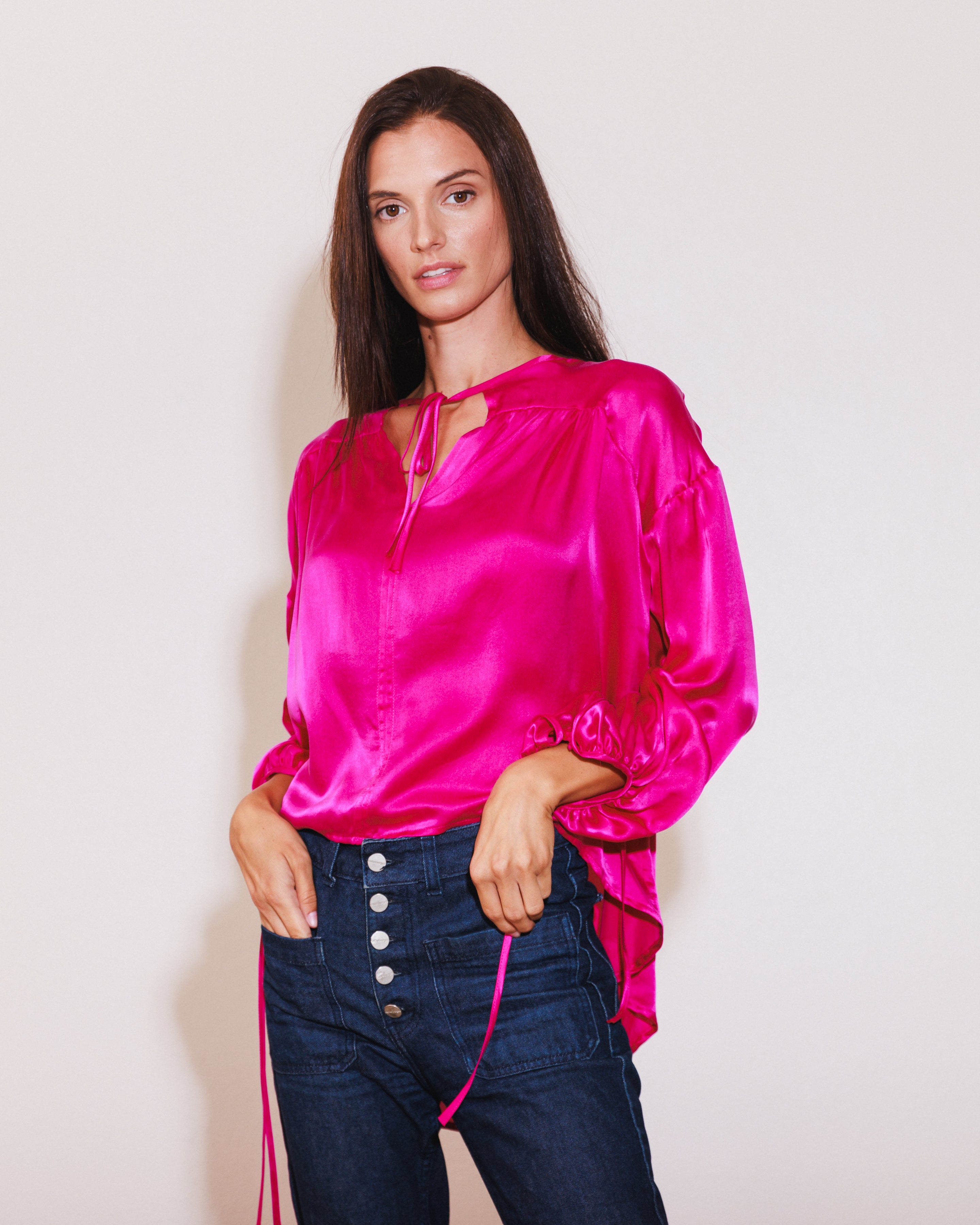 A woman wearing a Catherine Gee Frances Hot Pink blouse made of a shiny, satin-like fabric and features a vibrant hot pink color paired with high-waisted dark blue jeans with multiple buttons. It has long sleeves with gathered cuffs and a keyhole neckline with a tie detail. The combination of the bright blouse and dark jeans creates a striking contrast, making the outfit visually appealing and stylish.