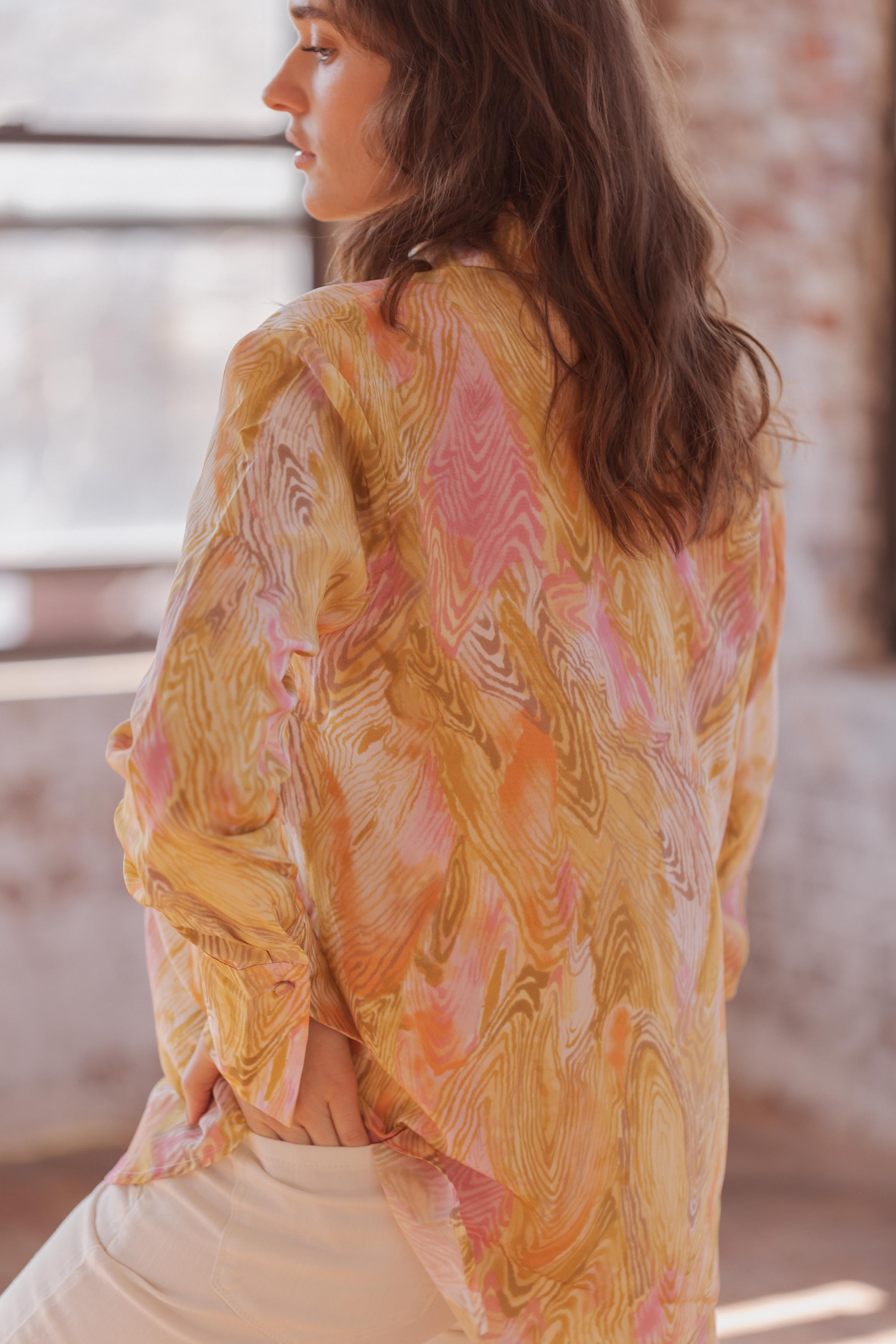 Back-view of a woman has one hand in a pocket wearing Catherine Gee Daria French Cuff Silk Topo Print blouse features a vibrant pattern with swirls of pink, yellow, and beige, creating a dynamic and eye-catching design. The fabric appears lightweight and flowy, contributing to a relaxed and elegant look, that adds a casual touch to the sophisticated garment.