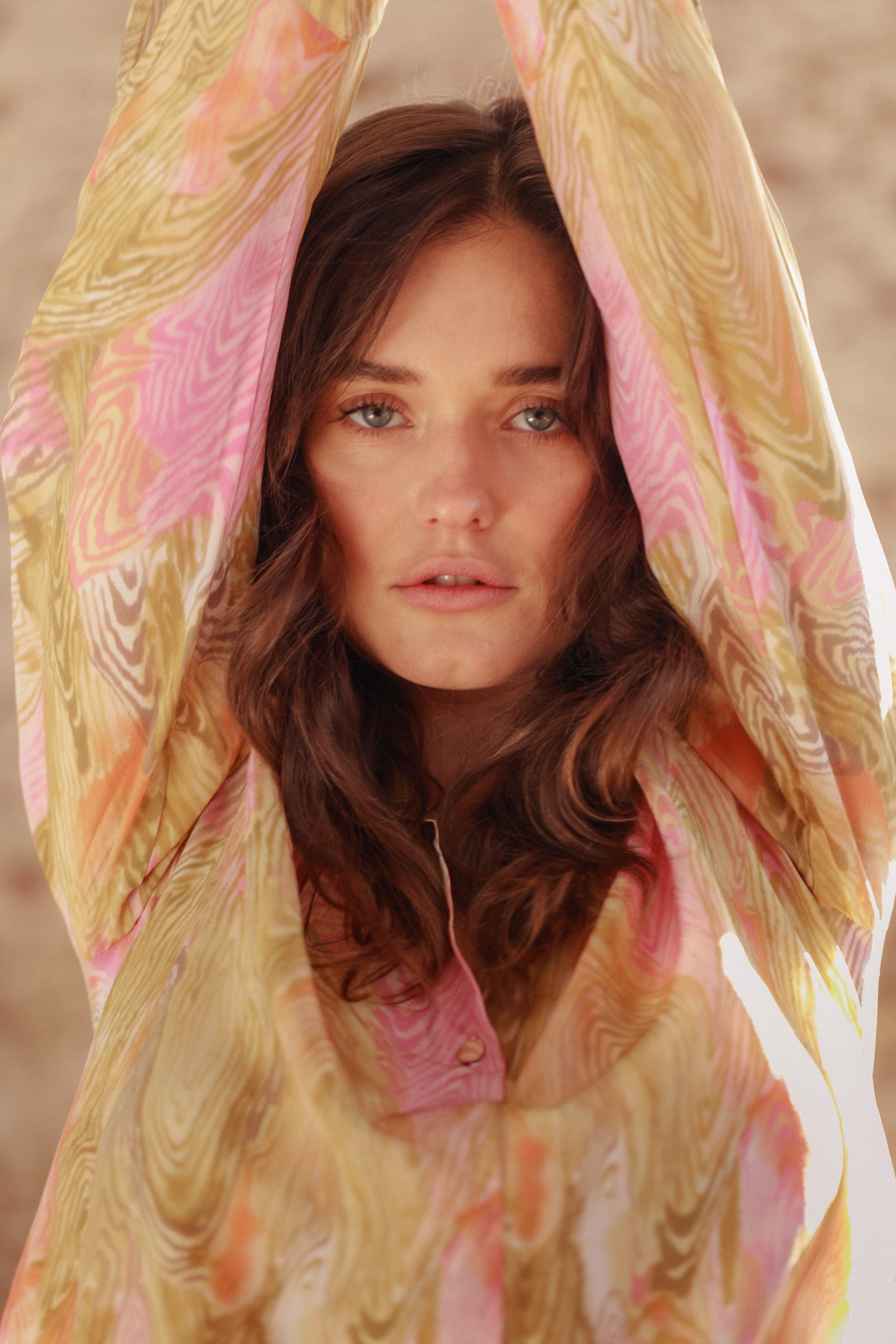 A woman has her arms raised wearing a Catherine Gee Daria French Cuff Silk Topo Print blouse that features a vibrant and intricate pattern with swirls of pink, yellow, and beige colors. The fabric appears to be lightweight and silky, draping elegantly, highlighting the long sleeves and the flowy nature of the blouse making the design and colors of the blouse visually striking and fashionable.