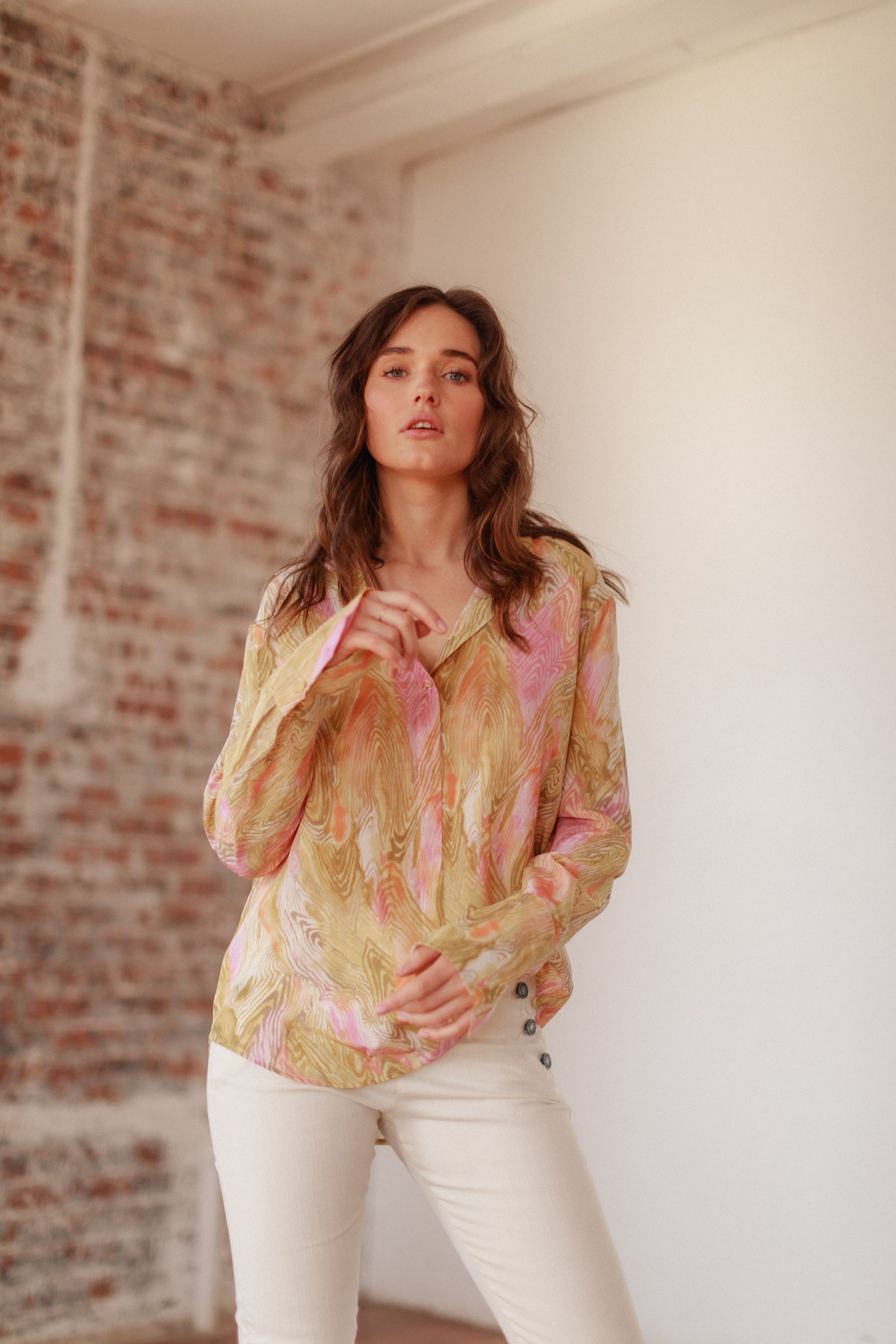 A woman wearing a Catherine Gee Daria French Cuff Silk Topo Print blouse features a colorful, abstract pattern with shades of pink, yellow, and beige paired with light-colored pants with button details on the side. The fabric appears lightweight and silky, with a loose fit and long sleeves ending in French cuffs. The background includes a brick wall and a white wall, adding a contrasting texture and highlighting the elegant look of the garment.