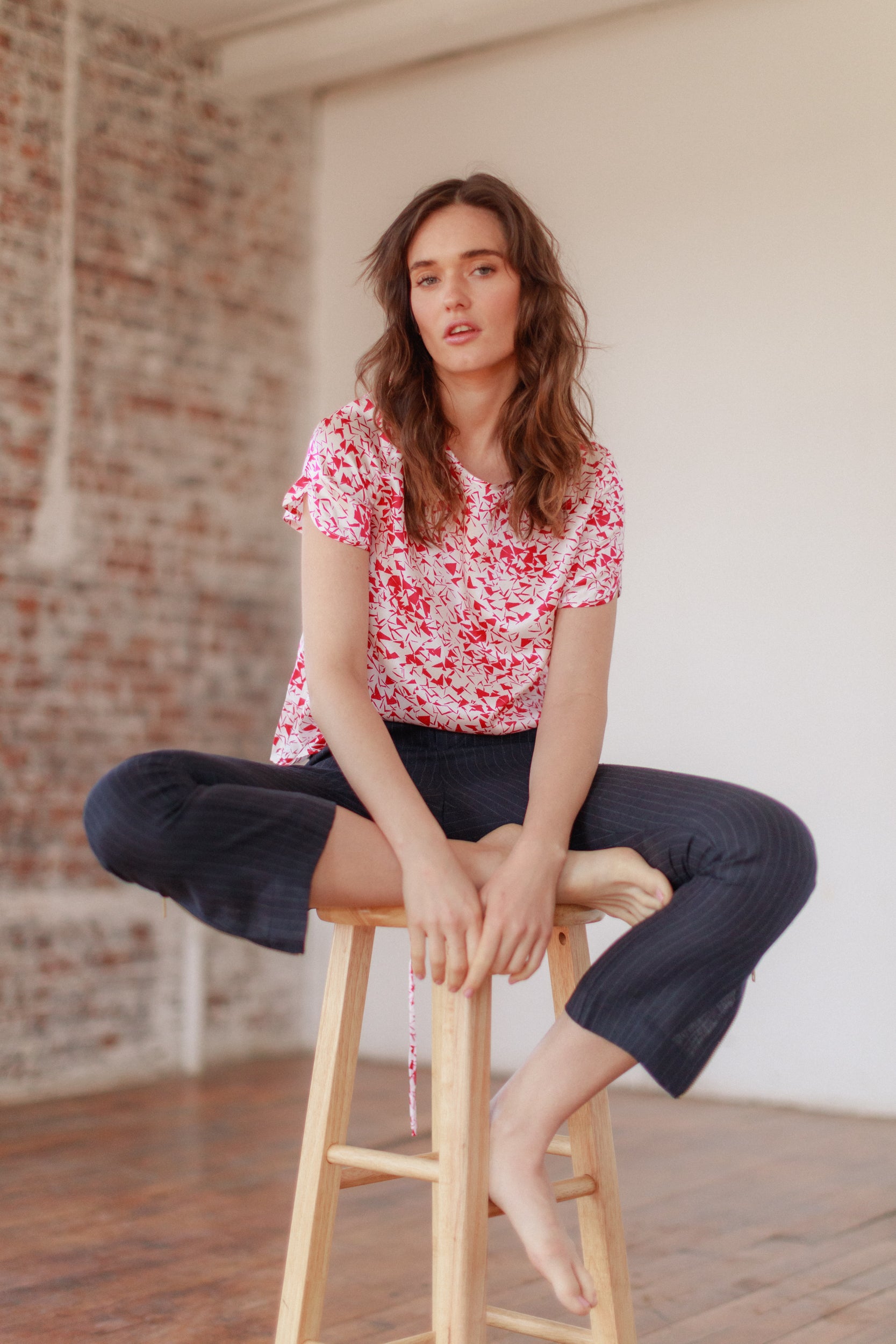 A woman is sitting cross-legged on a wooden stool in a room with a brick wall and wooden floor wearing a pink and white floral-patterned shirt and dark pants. The Catherine Gee Cate Ruched Back Shirt.