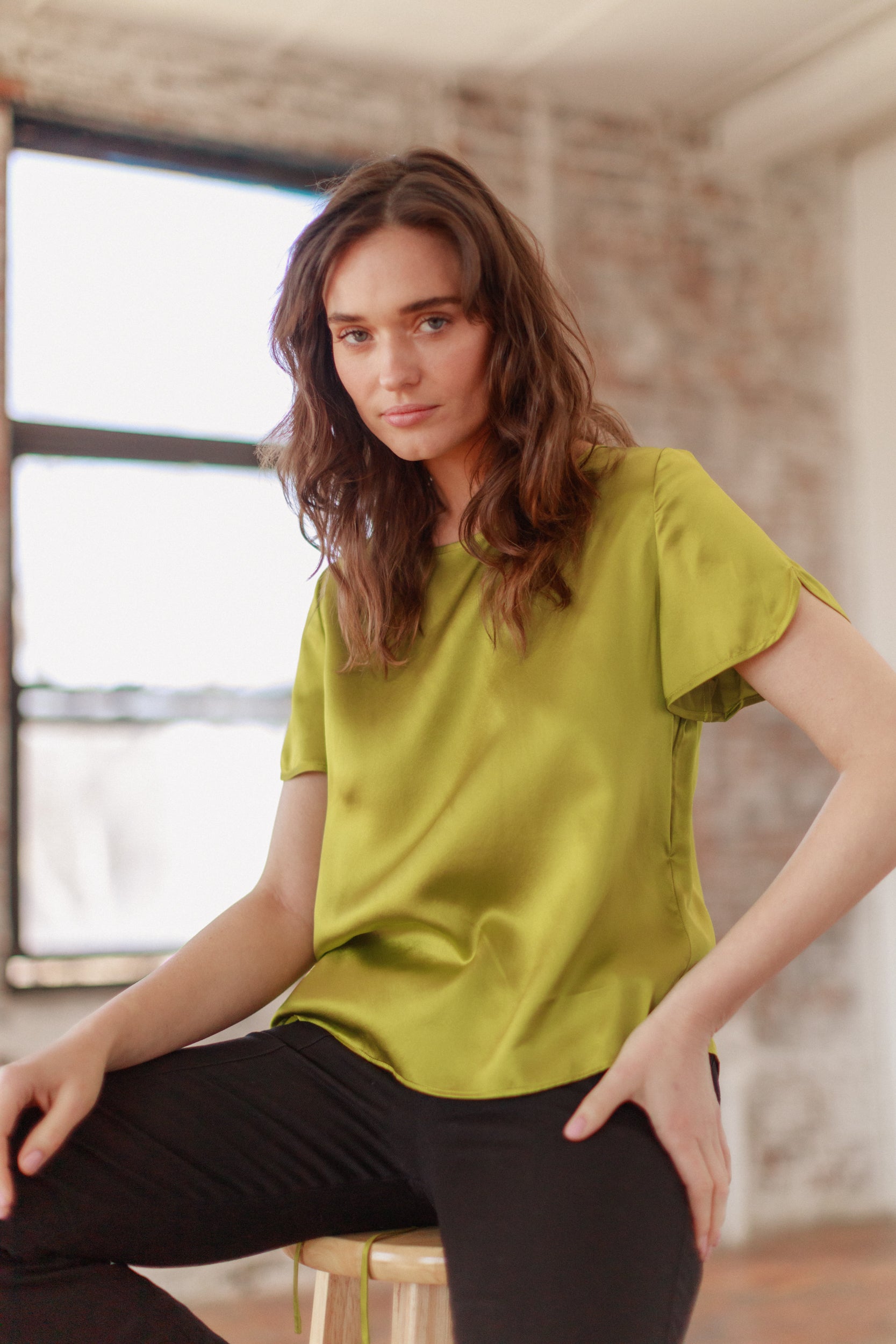 A woman is wearing a Catherine Gee Cate Ruched Back Shirt in a vibrant green color. The shirt has short sleeves and a smooth, silky texture. She is seated on a stool with one hand resting on the knee and the other on the stool. The background features a large window with natural light coming through and a brick wall, giving the setting a casual and relaxed atmosphere.