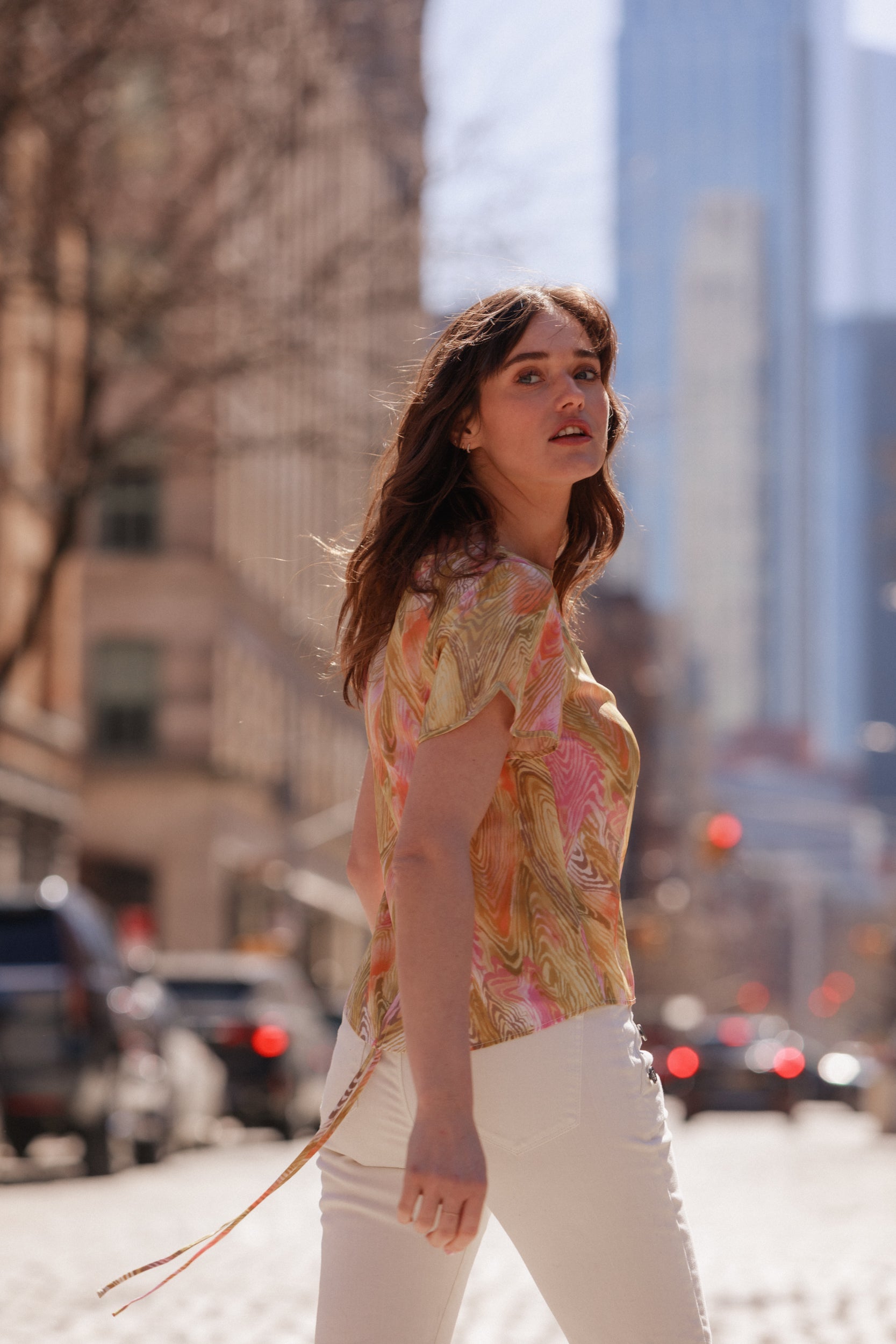 A woman is standing on a cobblestone street in an urban setting with tall buildings in the background. She is wearing a colorful, patterned short-sleeve shirt and white pants. The shirt is identified as the Catherine Gee Cate Ruched Back Shirt. The image is taken during the daytime, and the background is slightly blurred, focusing on the person. The scene is interesting due to the contrast between the vibrant shirt and the urban environment.