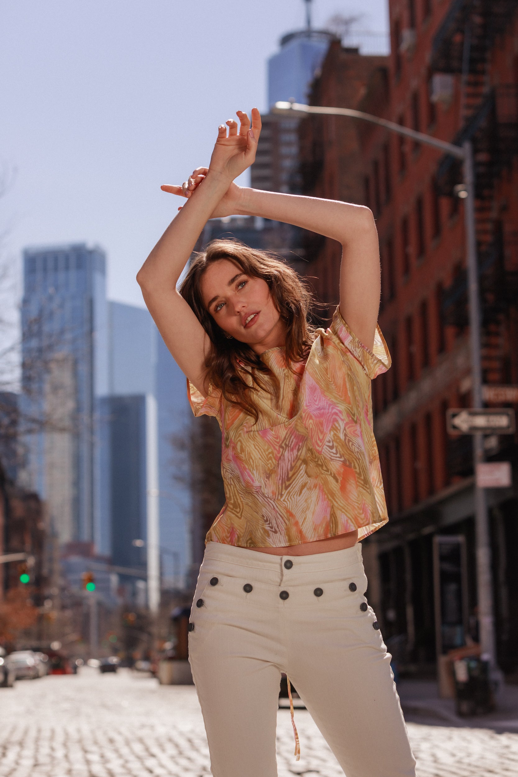 A woman standing on a cobblestone street, wearing Catherine Gee Josephine Nautical Skinny Pants. The high-waisted white pants feature button accents along the front, creating a chic nautical look paired with a vibrant, patterned top that adds a pop of color to the outfit. The background includes tall buildings, enhancing the urban vibe and highlighting the fashionable, dynamic nature of the ensemble.