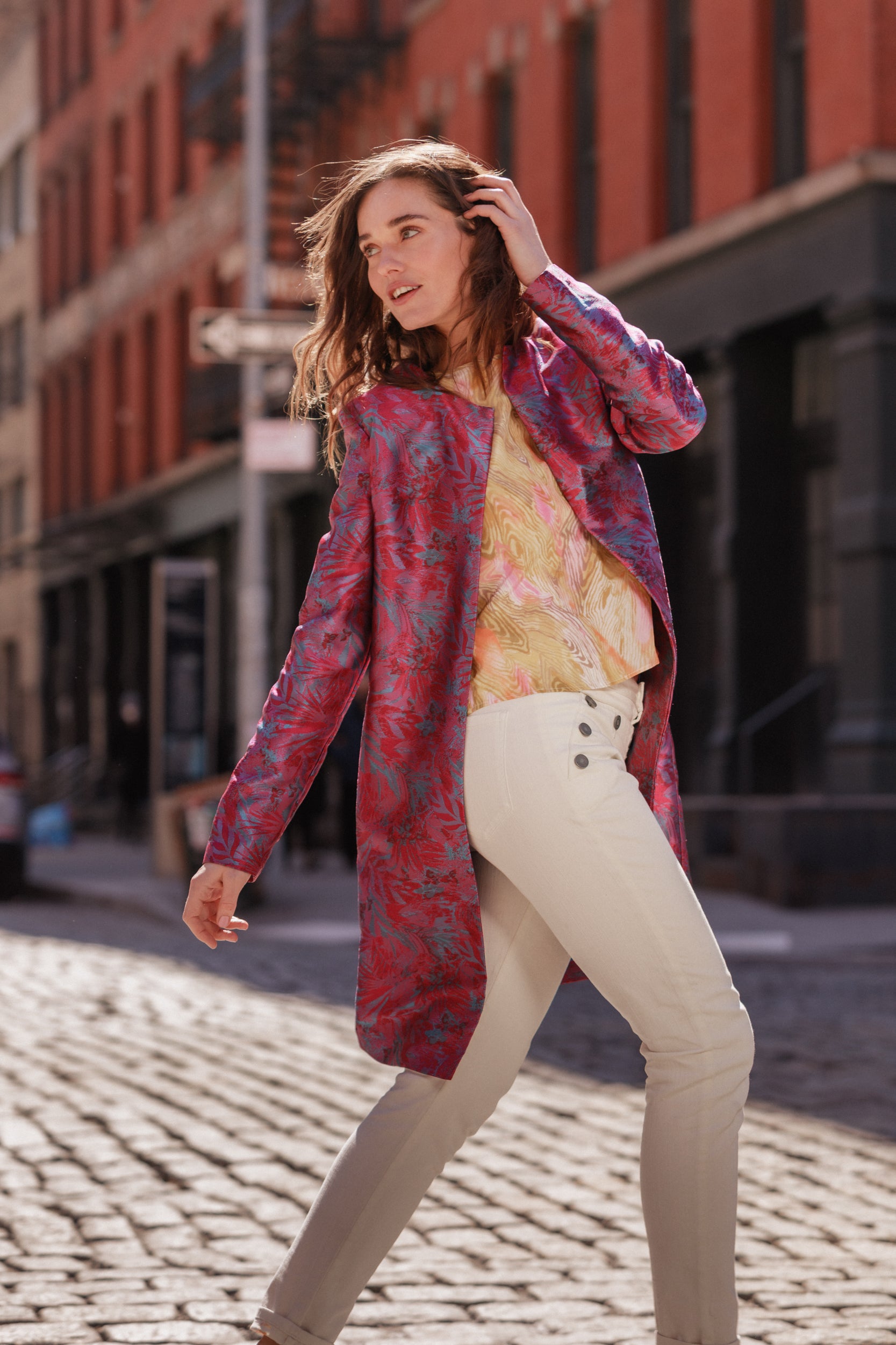 A woman walking on a cobblestone street in an urban setting, wearing Catherine Gee Josephine Nautical Skinny Pants. The high-waisted white pants feature distinctive button details along the front, creating a chic nautical look. She pairs them with a vibrant, patterned pink and purple long coat and a yellow top captures a fashionable street style, highlighting the pants as the key element of the getup.