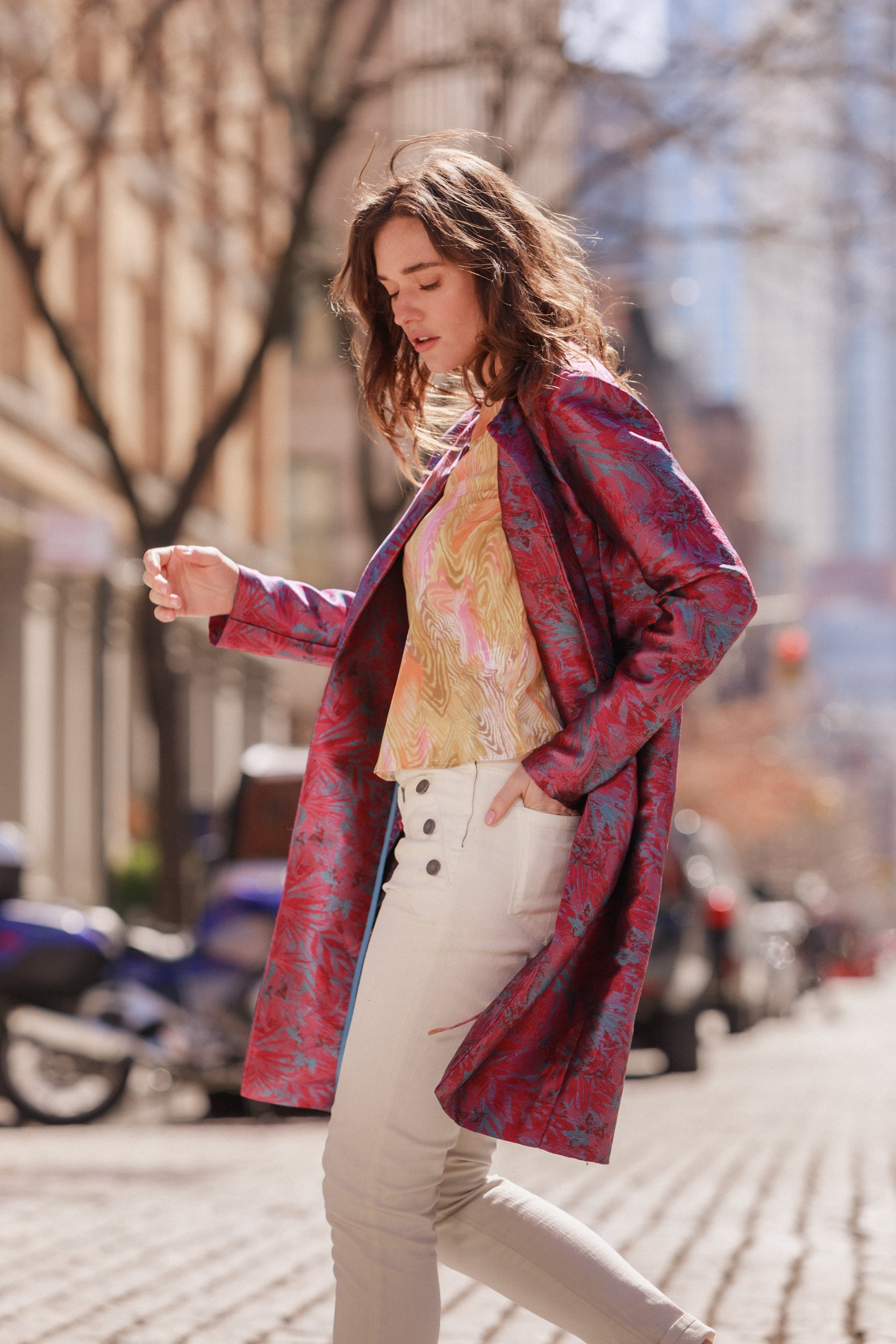 A woman walking on a cobblestone street, wearing Catherine Gee Josephine Nautical Skinny Pants. These high-waisted white pants feature stylish button accents on the front, creating a nautical-inspired look. She pairs them with a patterned pink and blue coat and a yellow and pink top. The urban background includes blurred buildings and vehicles, adding to the chic, city vibe of the outfit.