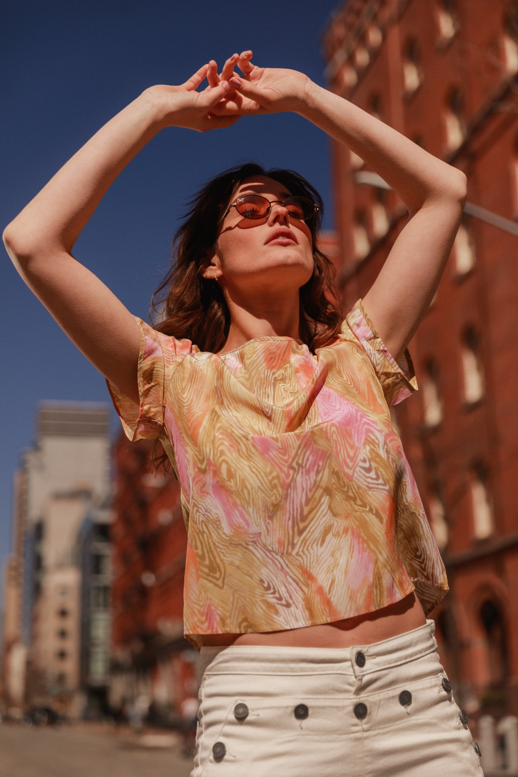 A woman wearing a Catherine Gee Cate Ruched Back Shirt stands outdoors with arms raised above her head. The shirt features a colorful, abstract pattern in shades of pink, yellow, and white. The person is also wearing high-waisted white pants with black buttons. The background shows a cityscape with red brick buildings and a clear blue sky.