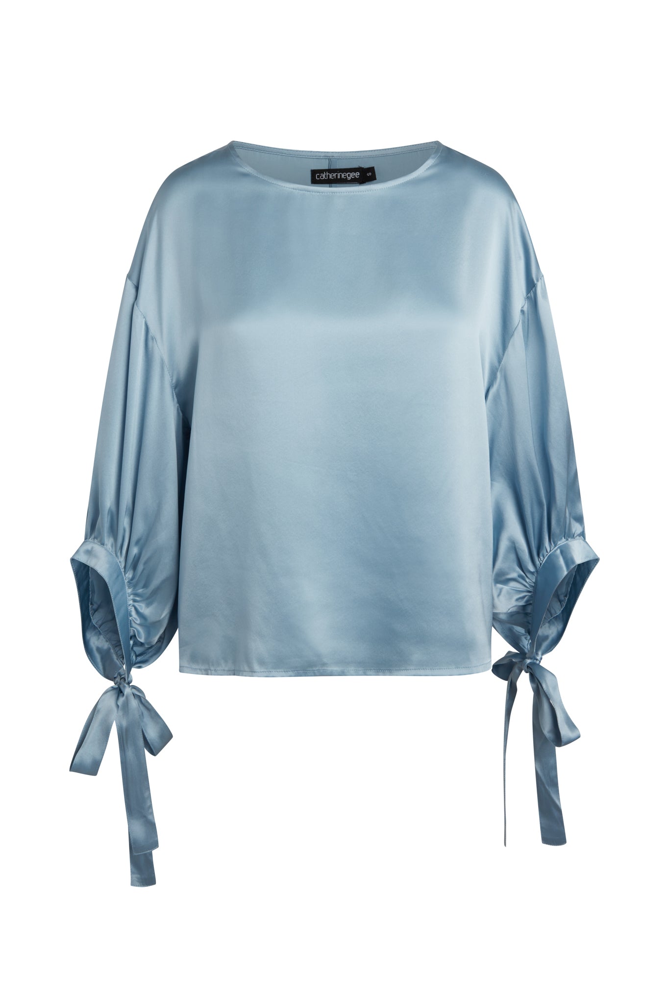 A periwinkle Catherine Gee Constance Blouse made of satin fabric features a loose, relaxed fit with a round neckline and three-quarter-length sleeves. The sleeves are gathered and tied with matching fabric ribbons at the cuffs, adding a stylish and elegant touch to the design.