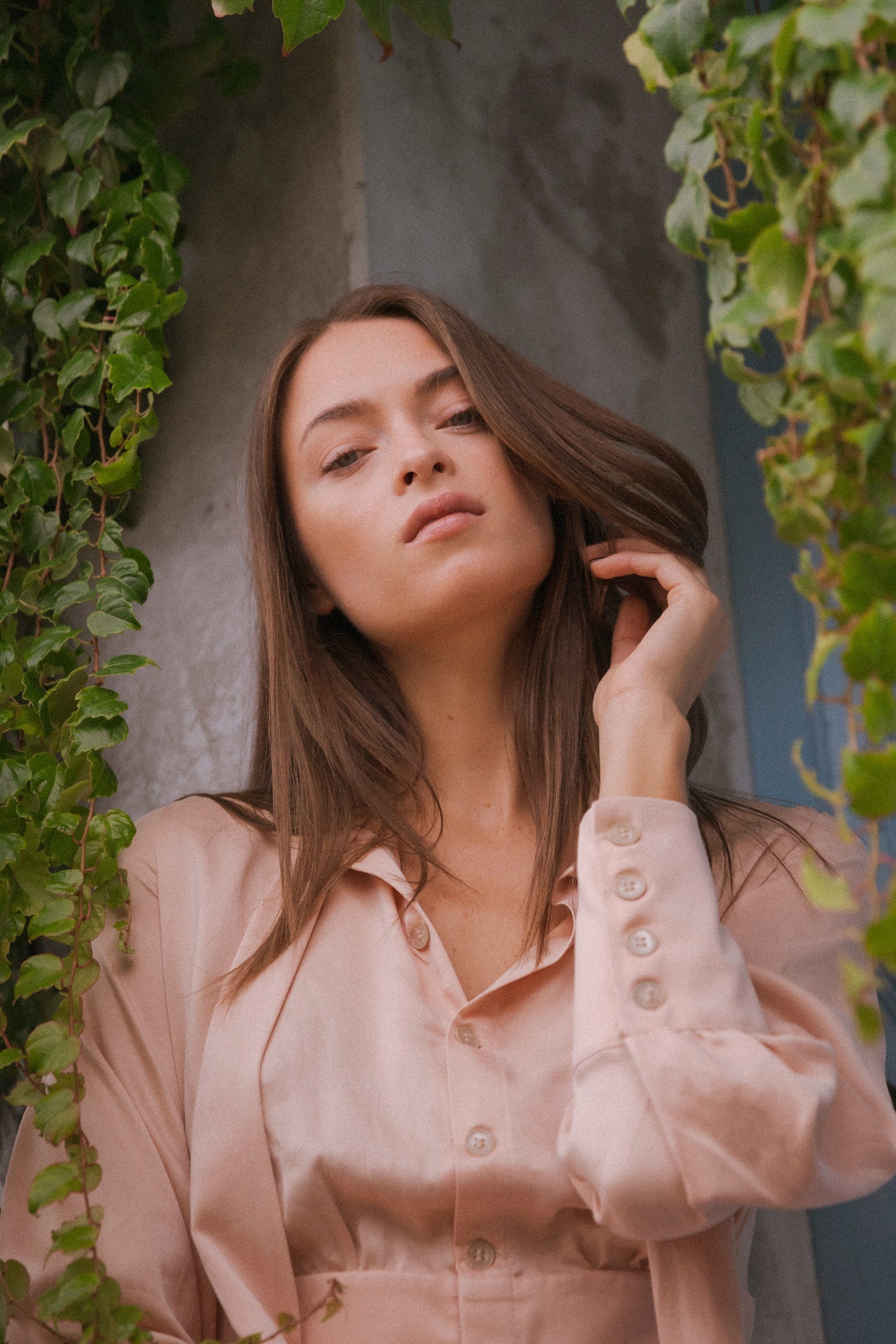 A woman standing amidst green foliage, holding a strand of her hair wearing a Catherine Gee Electric Nude color Flou High-Slit Long dress features long sleeves with buttoned cuffs, a high collar, and a fitted waist. The silky fabric flows elegantly, with a high slit that adds a touch of sophistication. The serene, natural setting, with a concrete wall and a blue door in the background, highlights the dress's luxurious design.