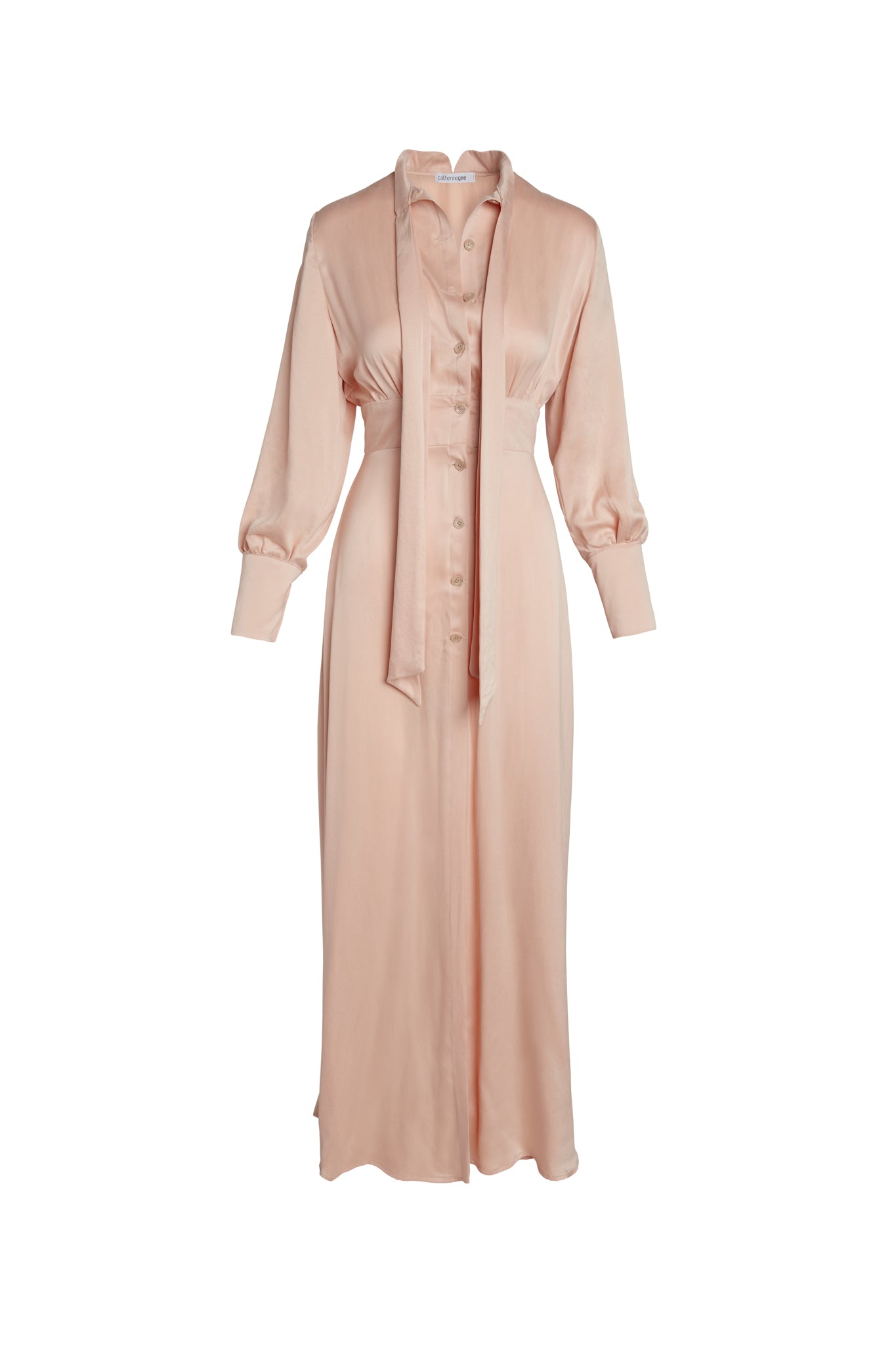 Catherine Gee Electric Flou High-Slit Long Dress. This elegant dress is made from a soft, flowing fabric in a Nude color and features a button-down front, long sleeves with cuffs, and a high slit that adds a touch of sophistication and allure. The dress also has a collar and a tie detail at the neckline, enhancing its chic and versatile design.