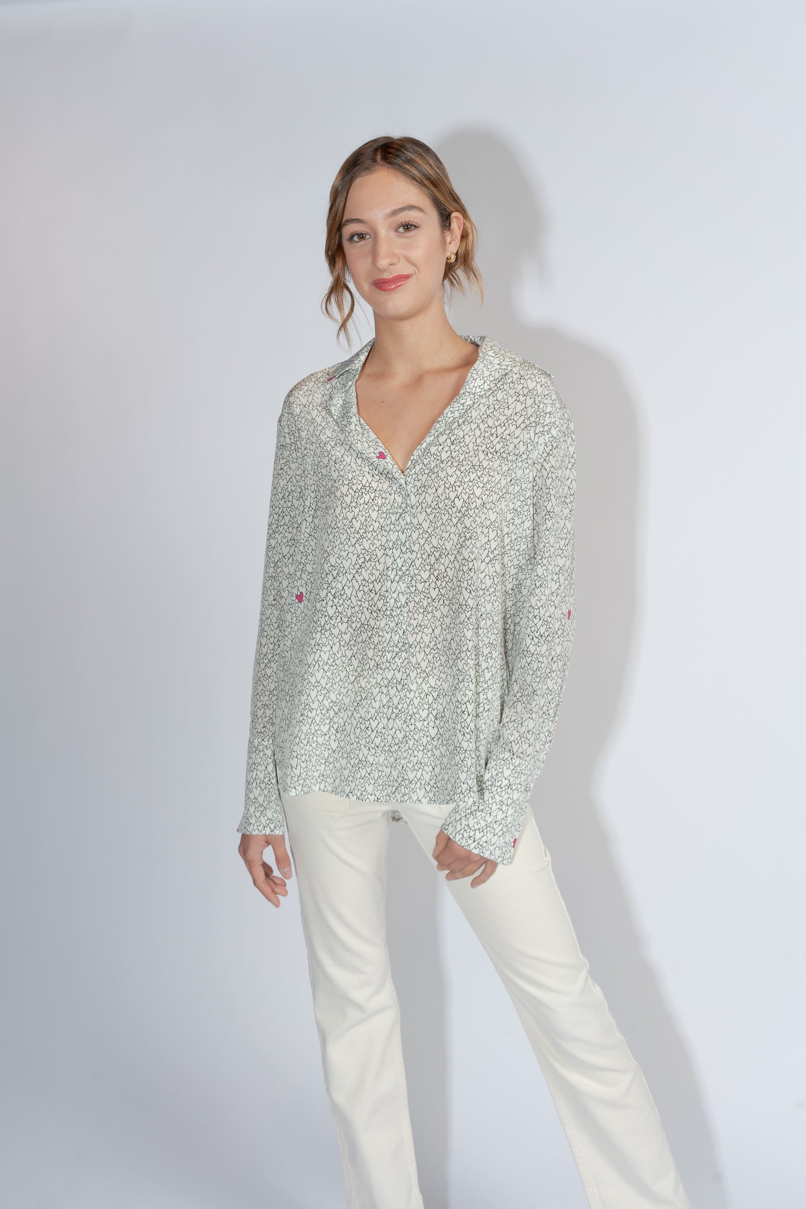 A woman wearing a Catherine Gee Daria French Cuff Silk-Heart Print blouse has a light, delicate heart print pattern and features a V-neckline paired with light-colored pants. The background is plain and light-colored, highlighting the outfit.